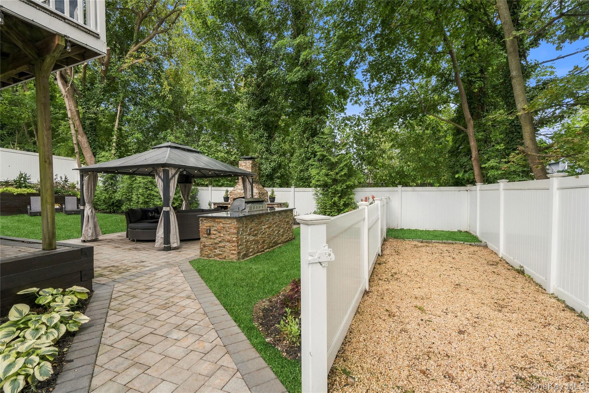 Backyard patio with a curtained gazebo, stone outdoor kitchen, lawn, and fenced gravel area.