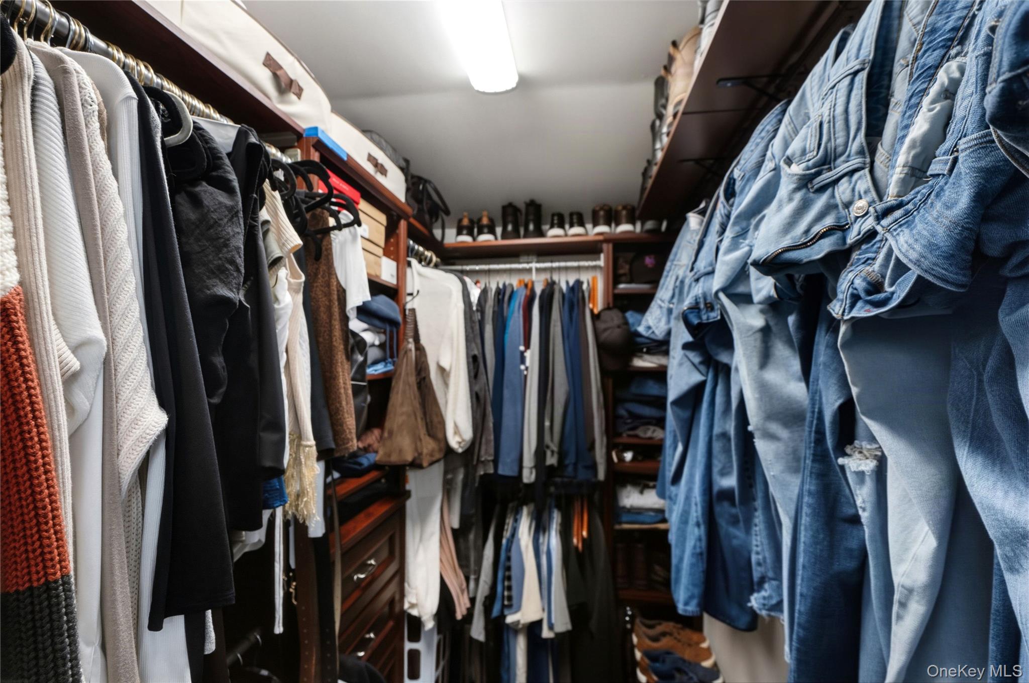 Walk-in closet with dark wood shelving, densely hung clothing, and shoes on display.