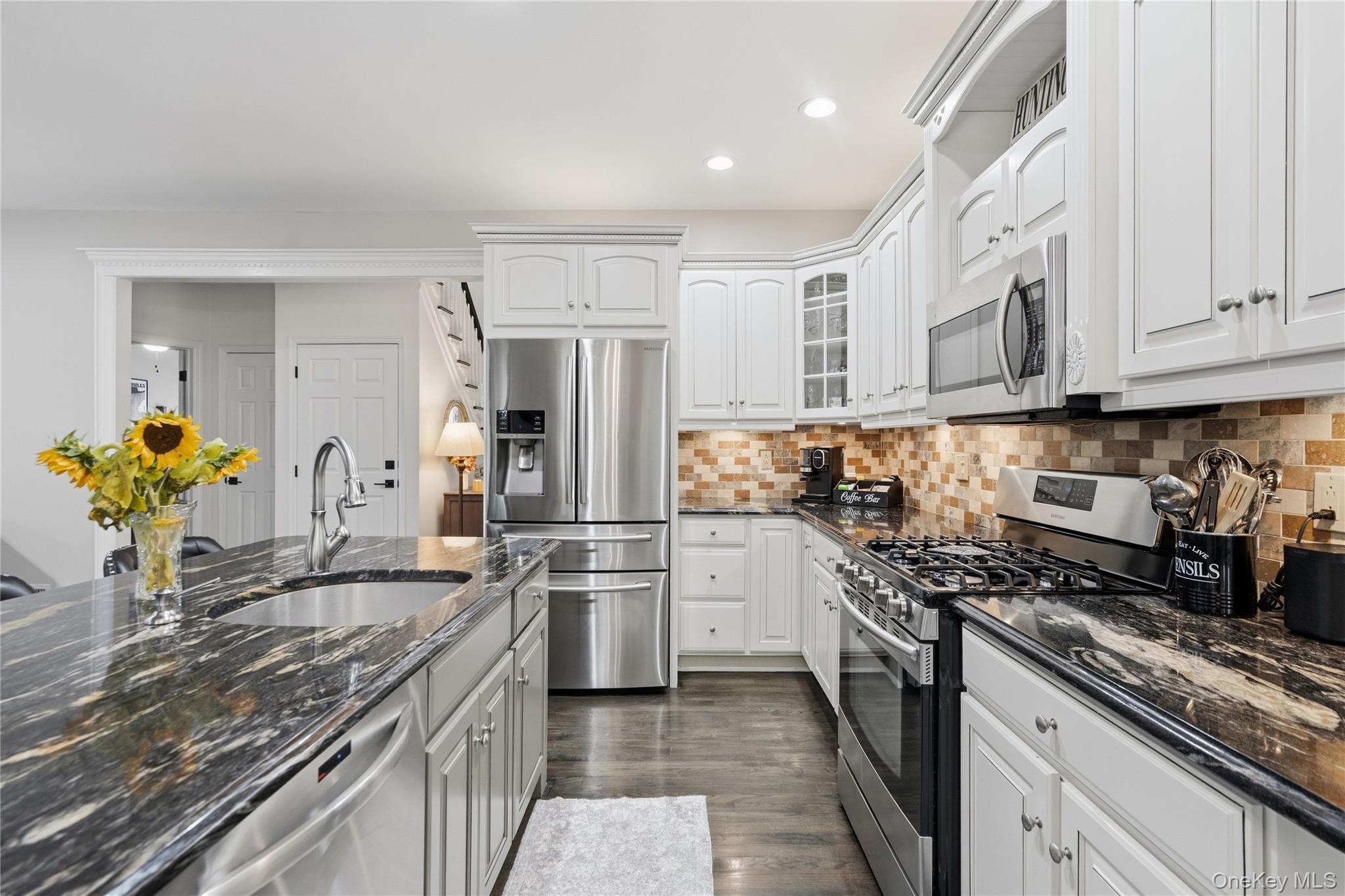 White kitchen with dark stone counters, stainless steel appliances, a tiled backsplash and sunflowers on the island.