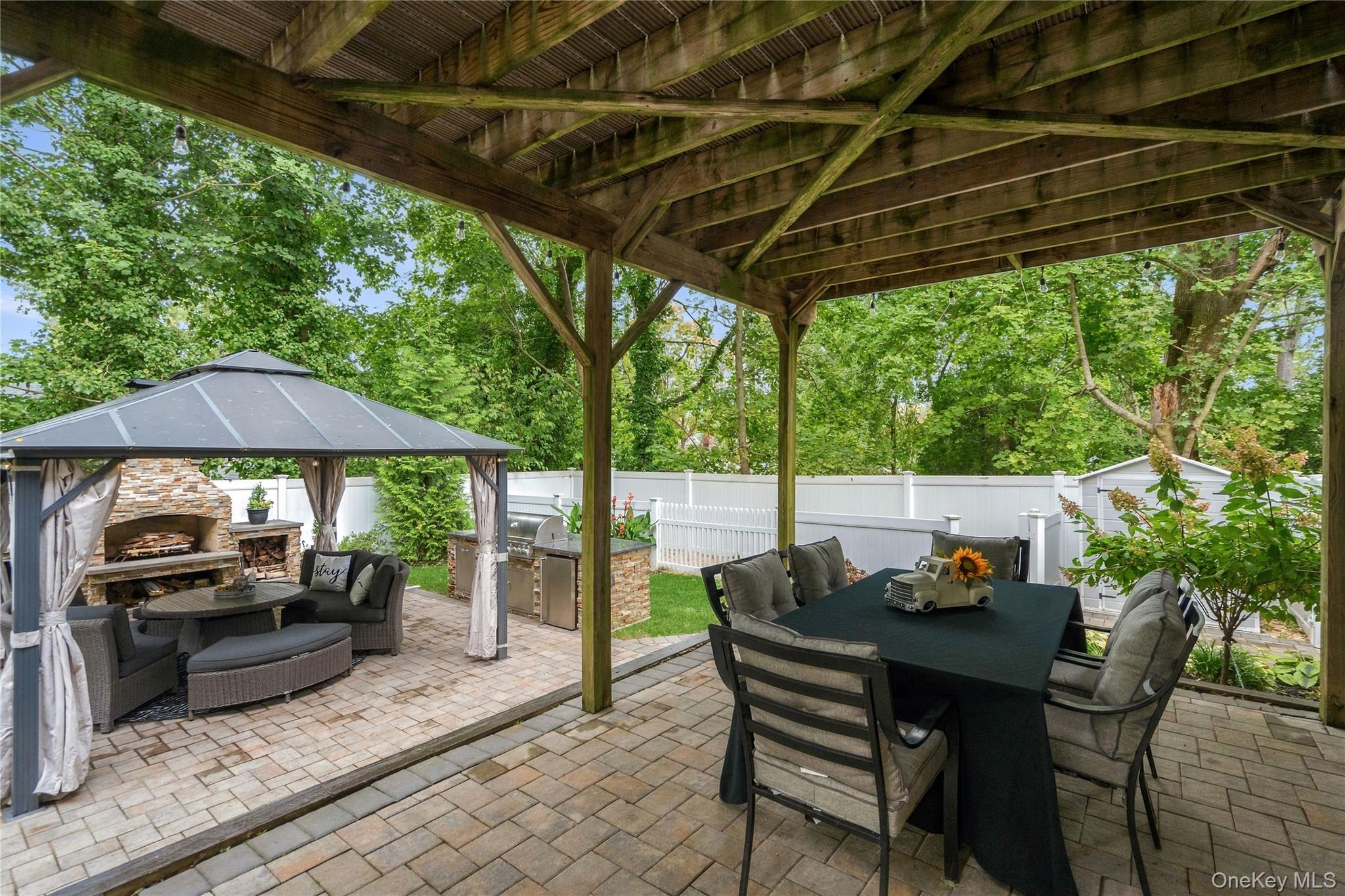 Furnished backyard patio under a timber pergola, with dining area and a separate seating nook by a stone fireplace.