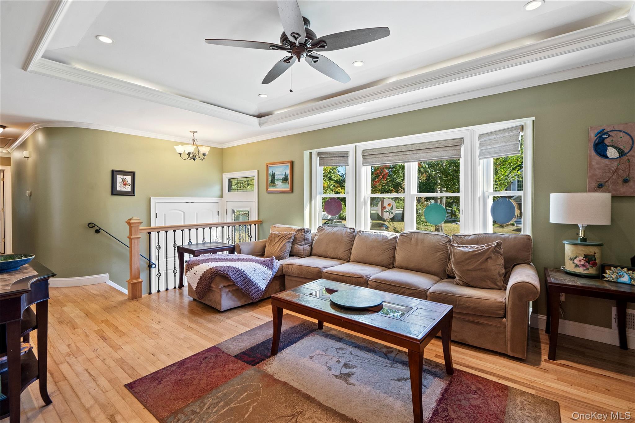 Living room with a brown sectional, hardwood floors, tray ceiling, and large windows overlooking trees.