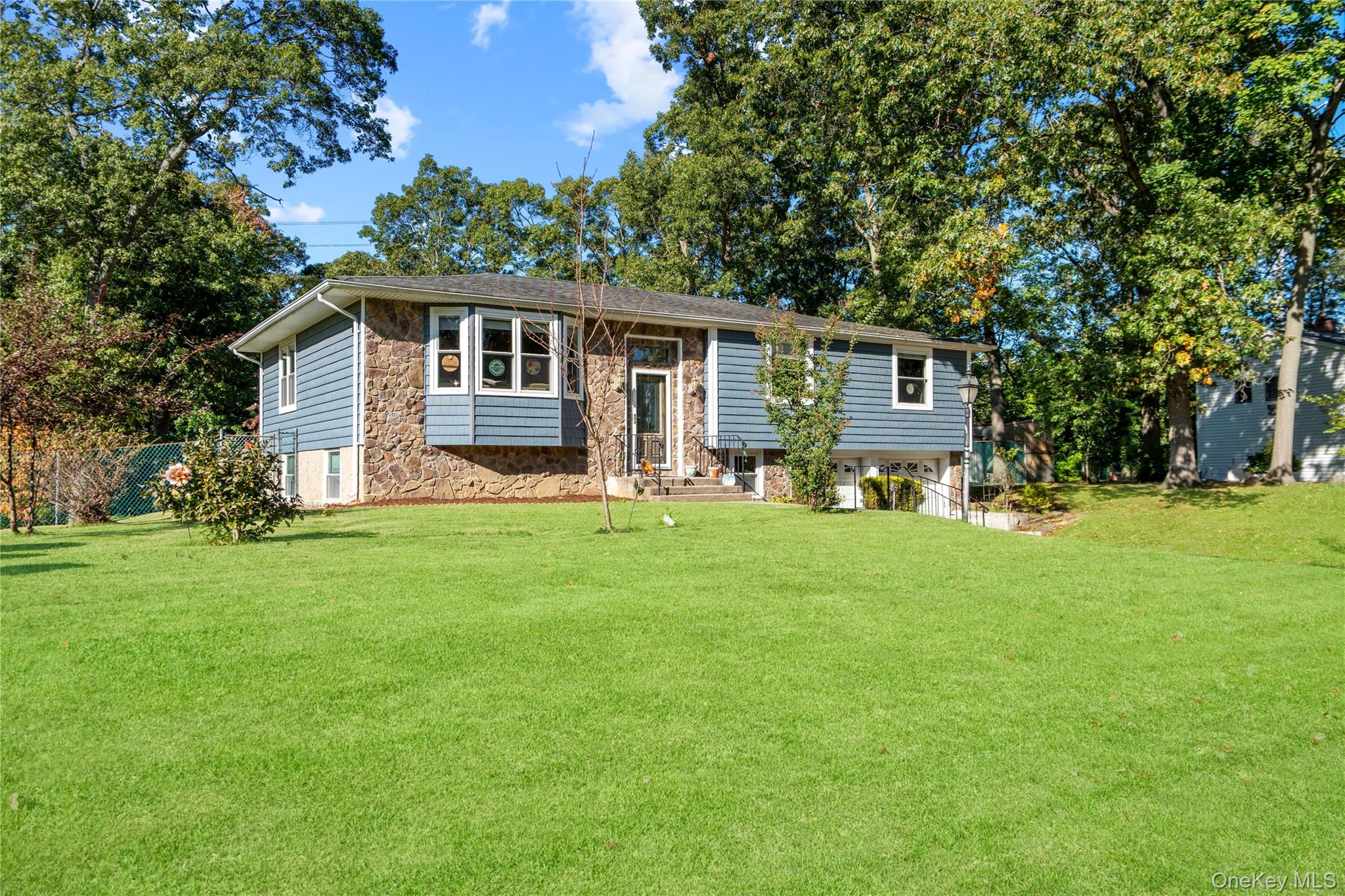 Split-level home with blue siding, stone facade, and a broad lawn beneath mature trees.