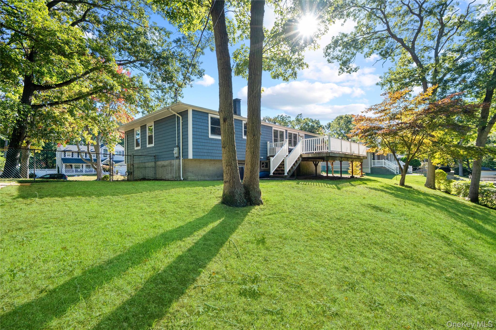 Rear of a blue-gray house with a raised white deck, framed by mature trees and a sunny lawn.