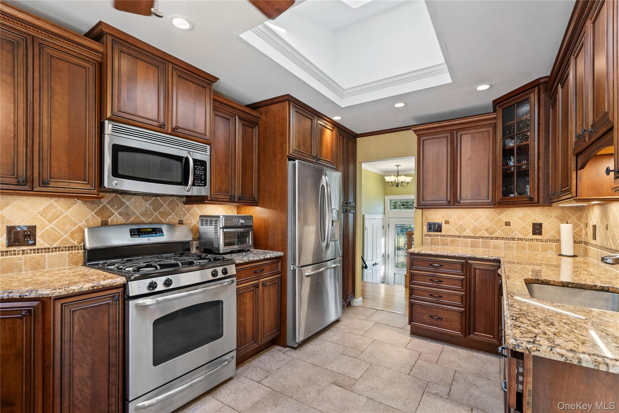 Kitchen with dark wood cabinetry, stainless steel appliances, tan stone counters, and a recessed tray ceiling.