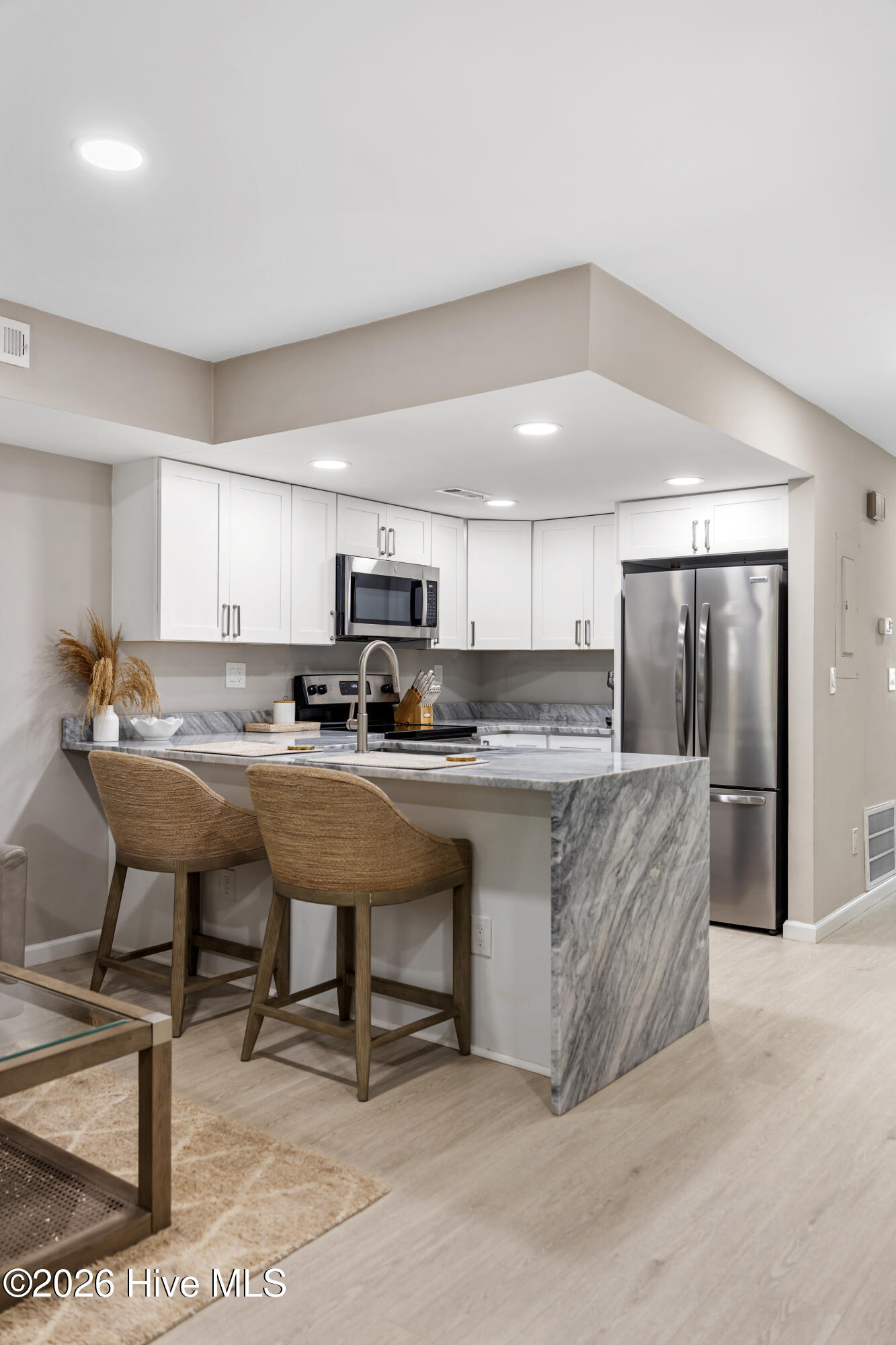 Staged kitchen with white cabinets, stainless appliances, and a marble-look peninsula with two woven stools.