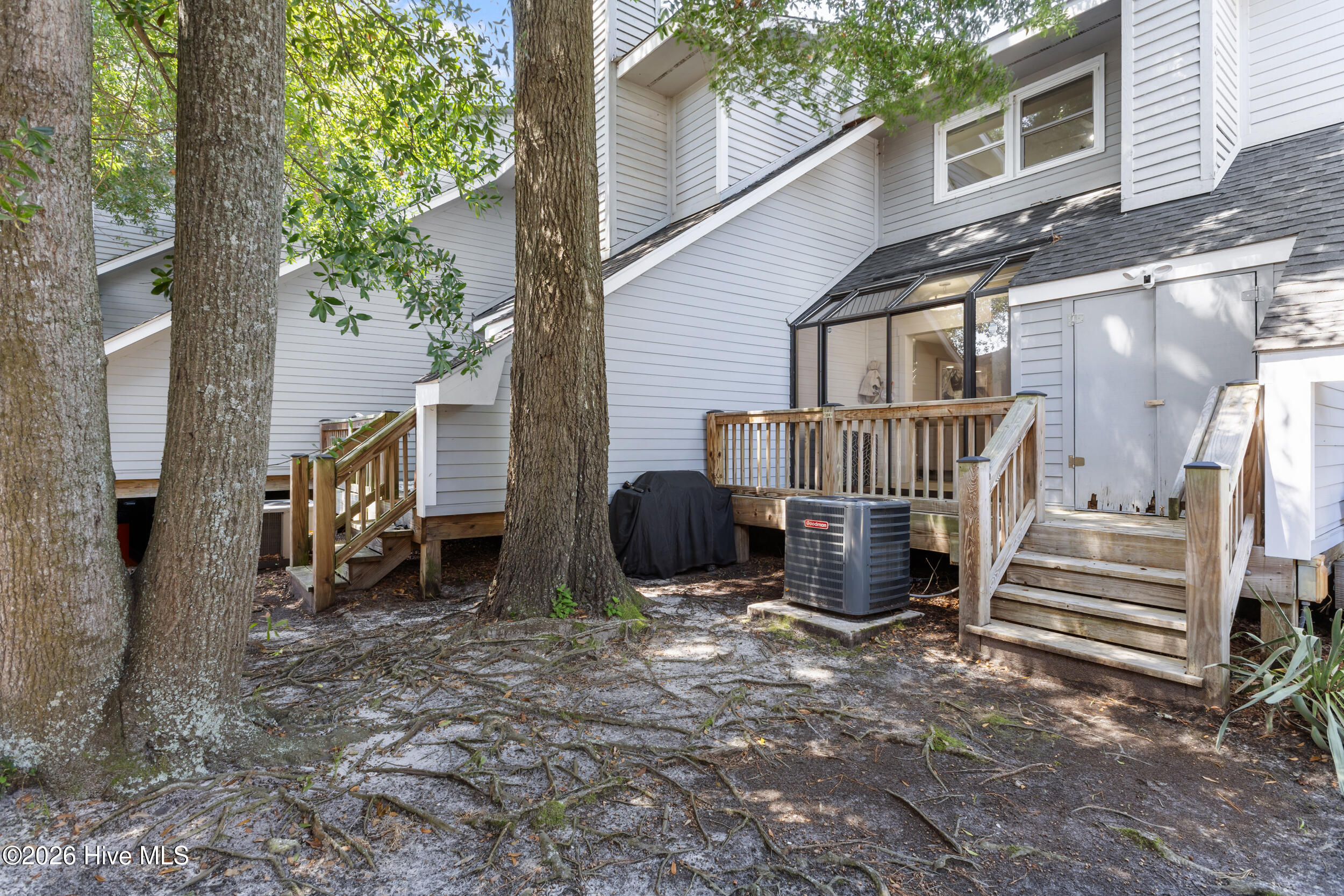 Home’s rear deck and stairs beside a glass-enclosed sunroom, shaded by mature trees.
