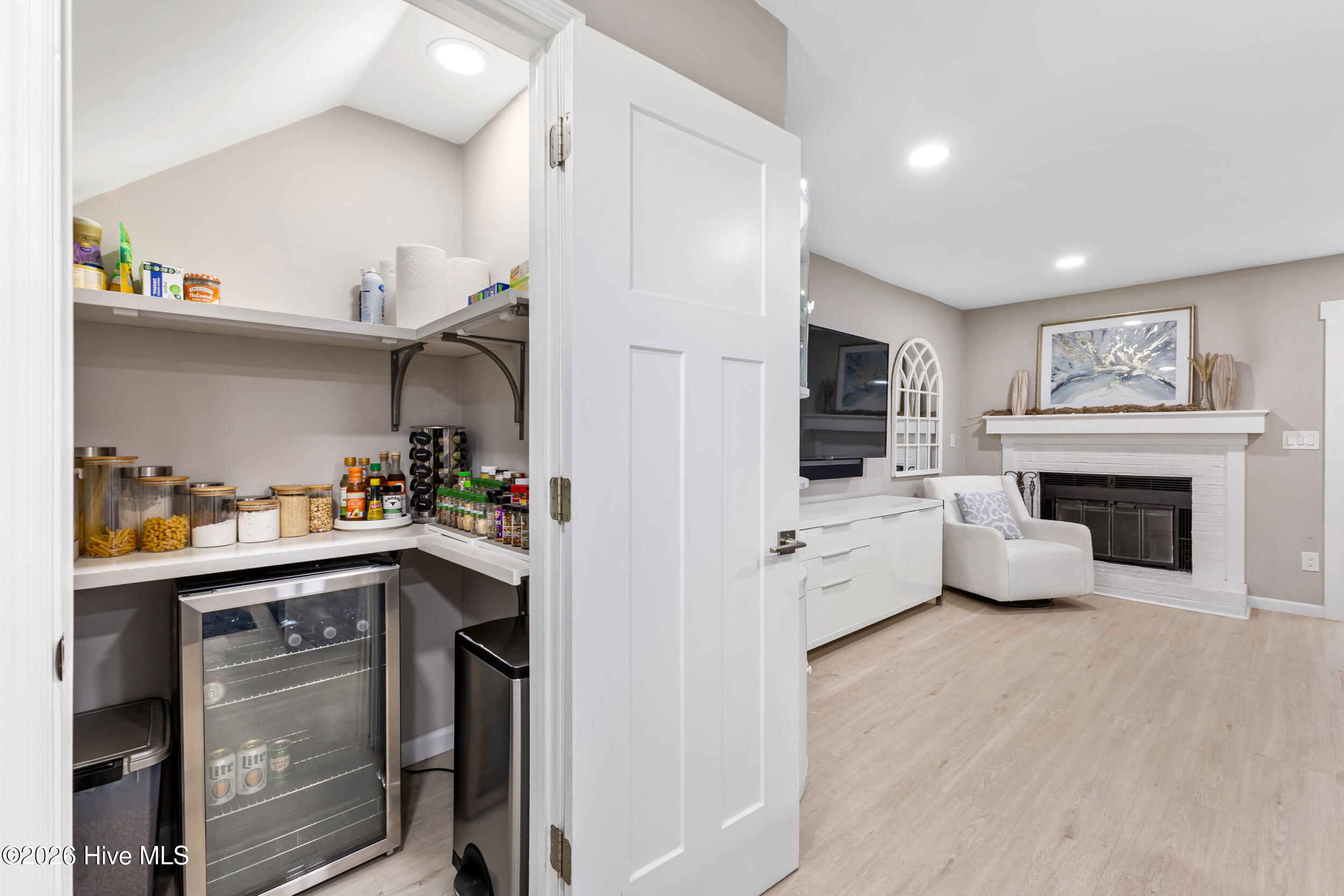 Stocked walk-in pantry with a glass-front beverage refrigerator opening onto a living room with a white fireplace.