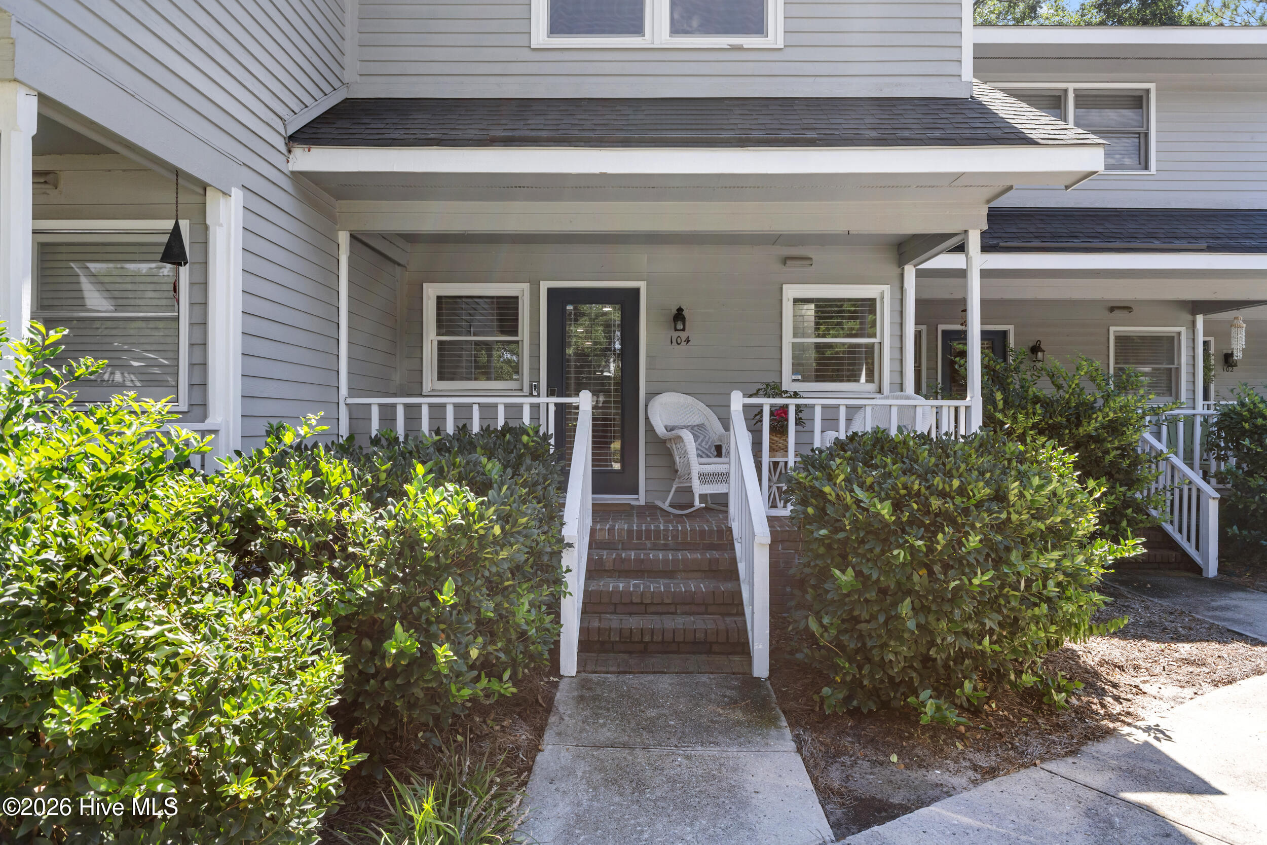 Unit 104 entrance with a covered porch, white railings, brick steps, and leafy shrubs.