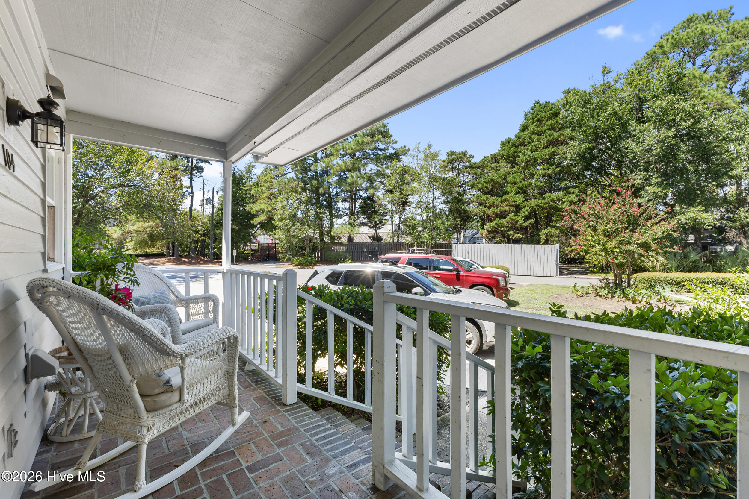 Covered front porch with white wicker rocking chairs, brick flooring, and a view of the landscaped yard.