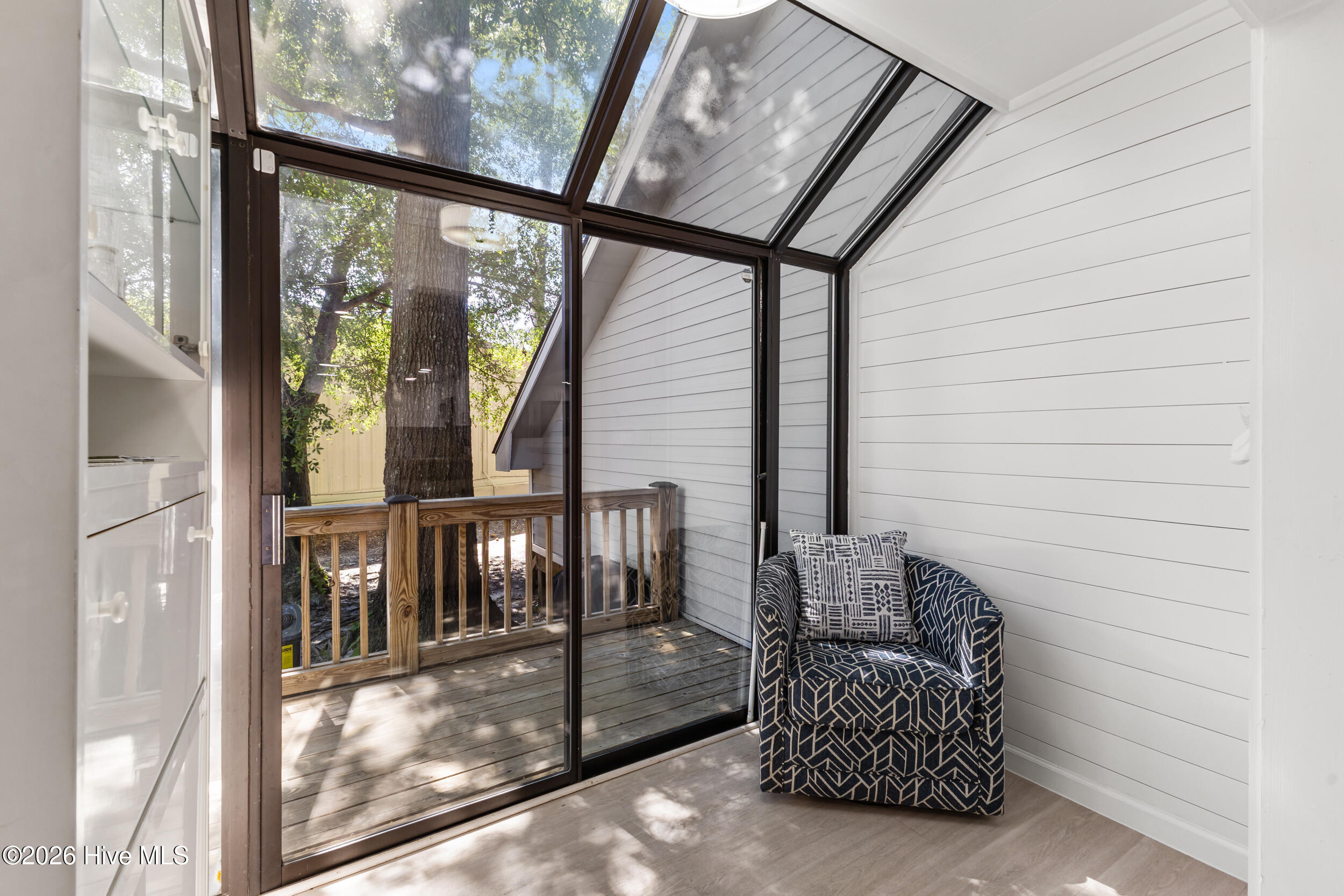 Sunroom with a patterned armchair, sloped glass ceiling, and views of a shaded wooden deck.
