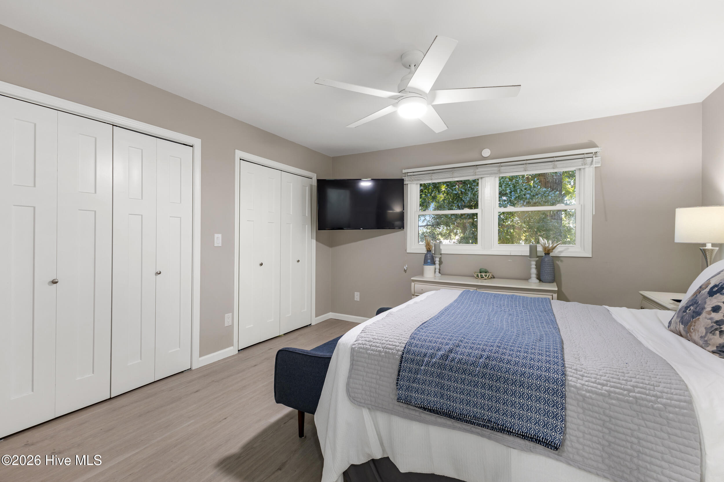 Bedroom with a blue patterned bed runner, leafy views through two windows, and a wall-mounted television.