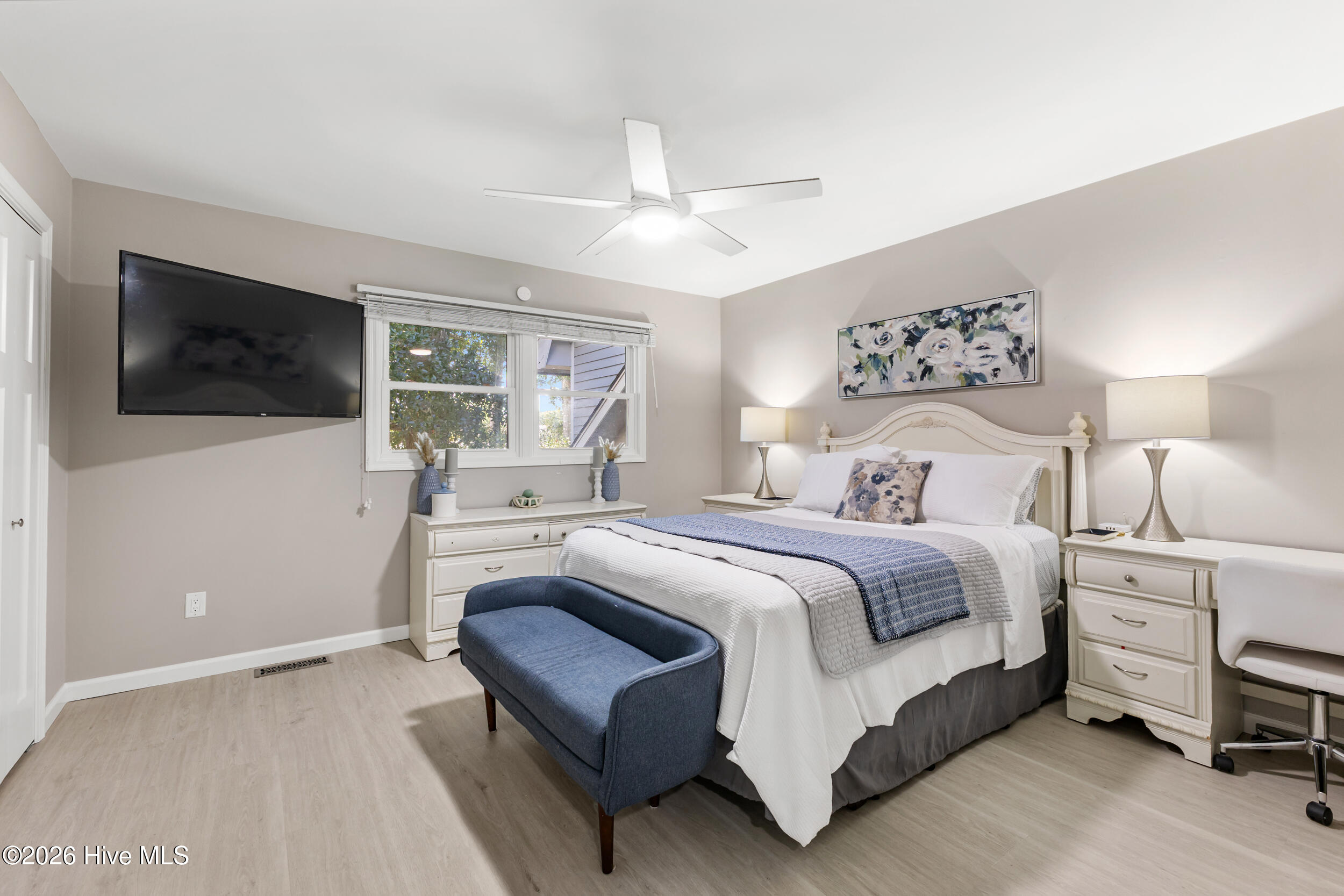 Furnished bedroom with a white bed, blue bench, matching nightstands, and a wall-mounted television.