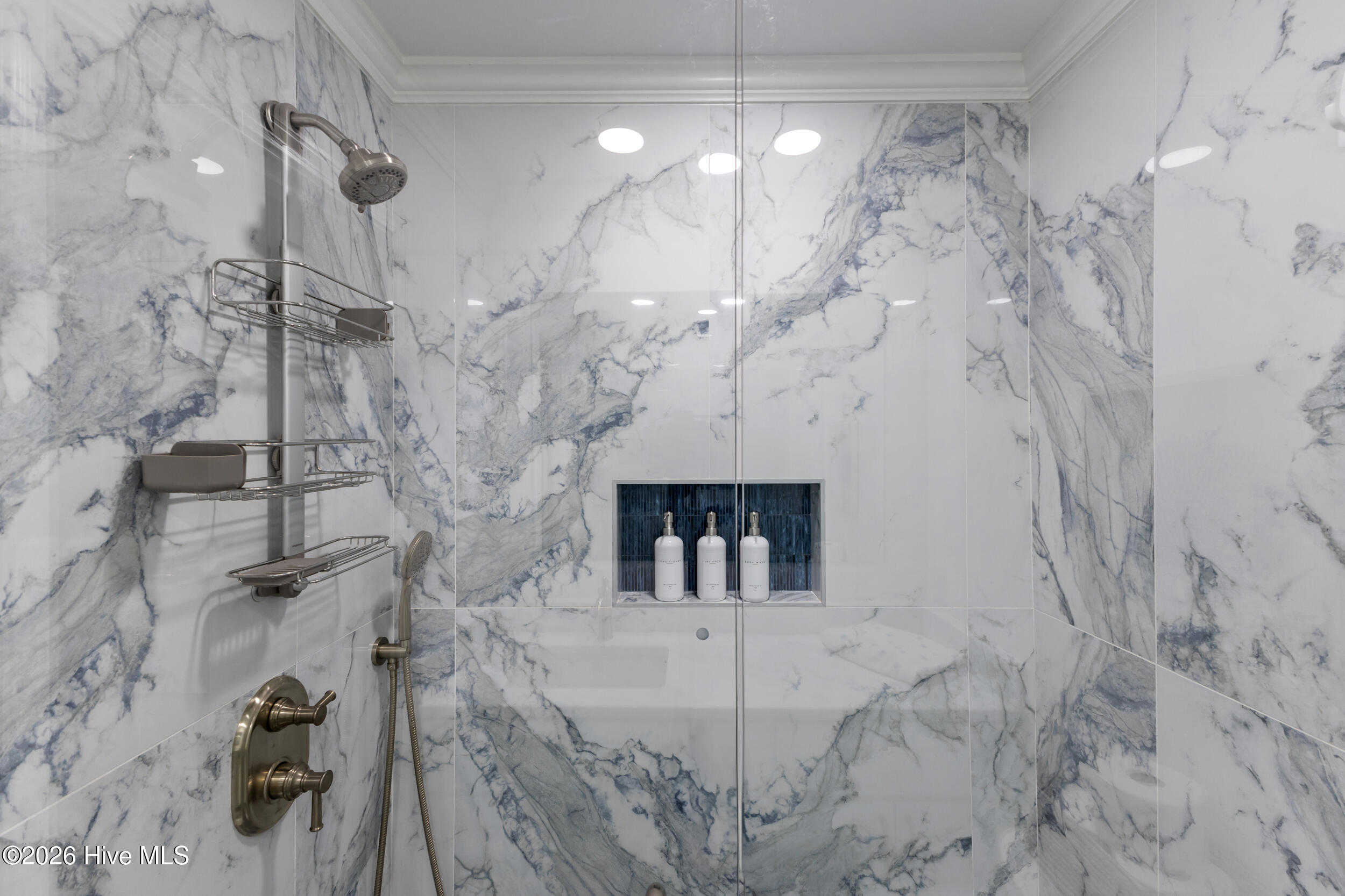 Marble-tiled shower with glass enclosure, metal fixtures, and recessed shelf of toiletries.