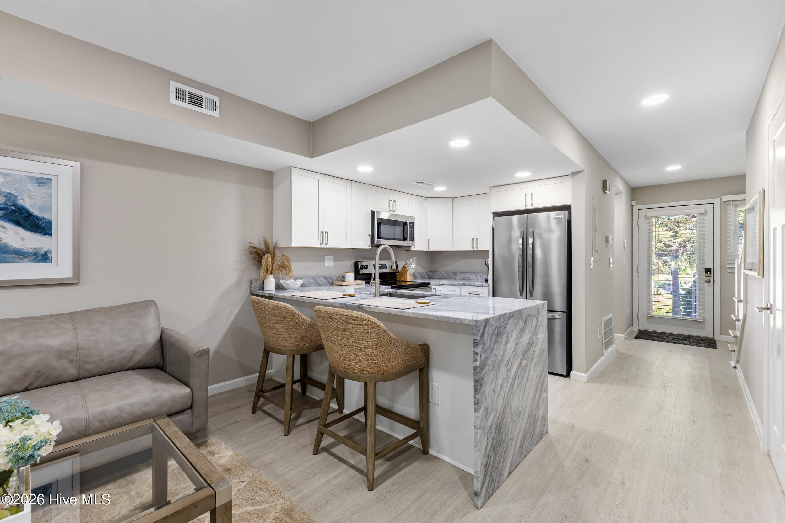 Open kitchen and living area with a marble-look island, woven stools, and stainless steel appliances.