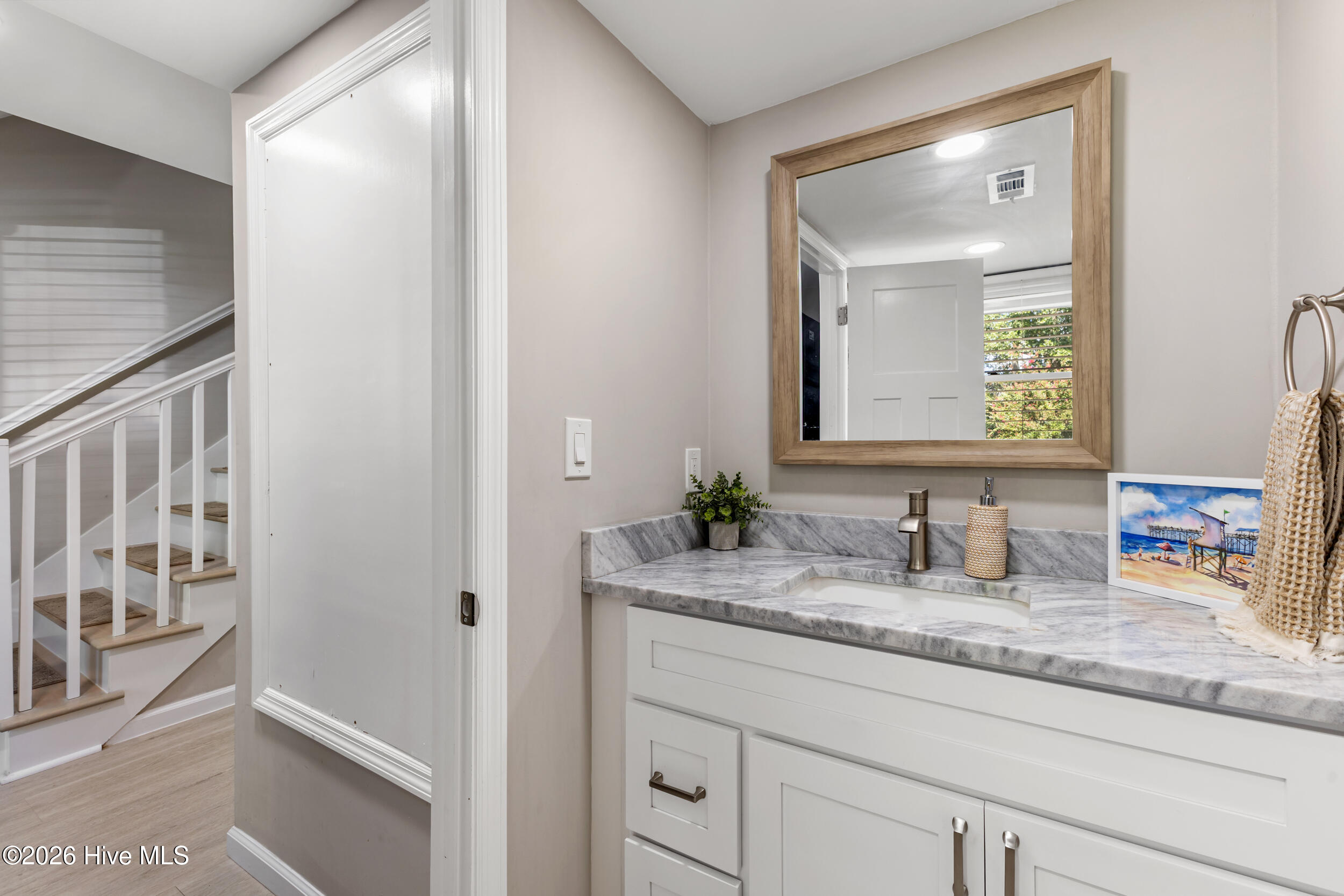 Bathroom vanity with gray marble counter and wood-framed mirror beside a staircase.
