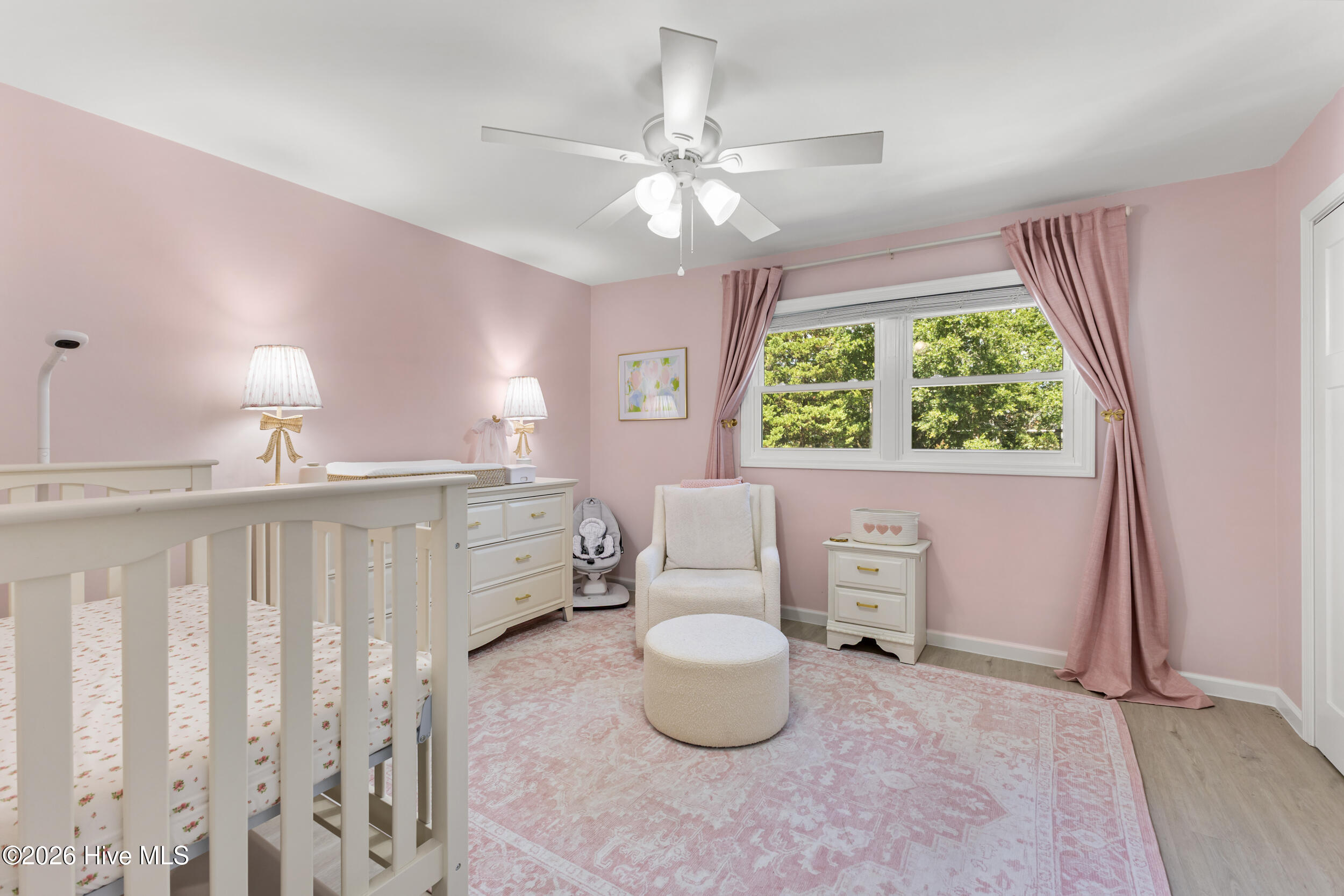 Nursery with a white crib, cream armchair, pink curtains, and a large window overlooking trees.