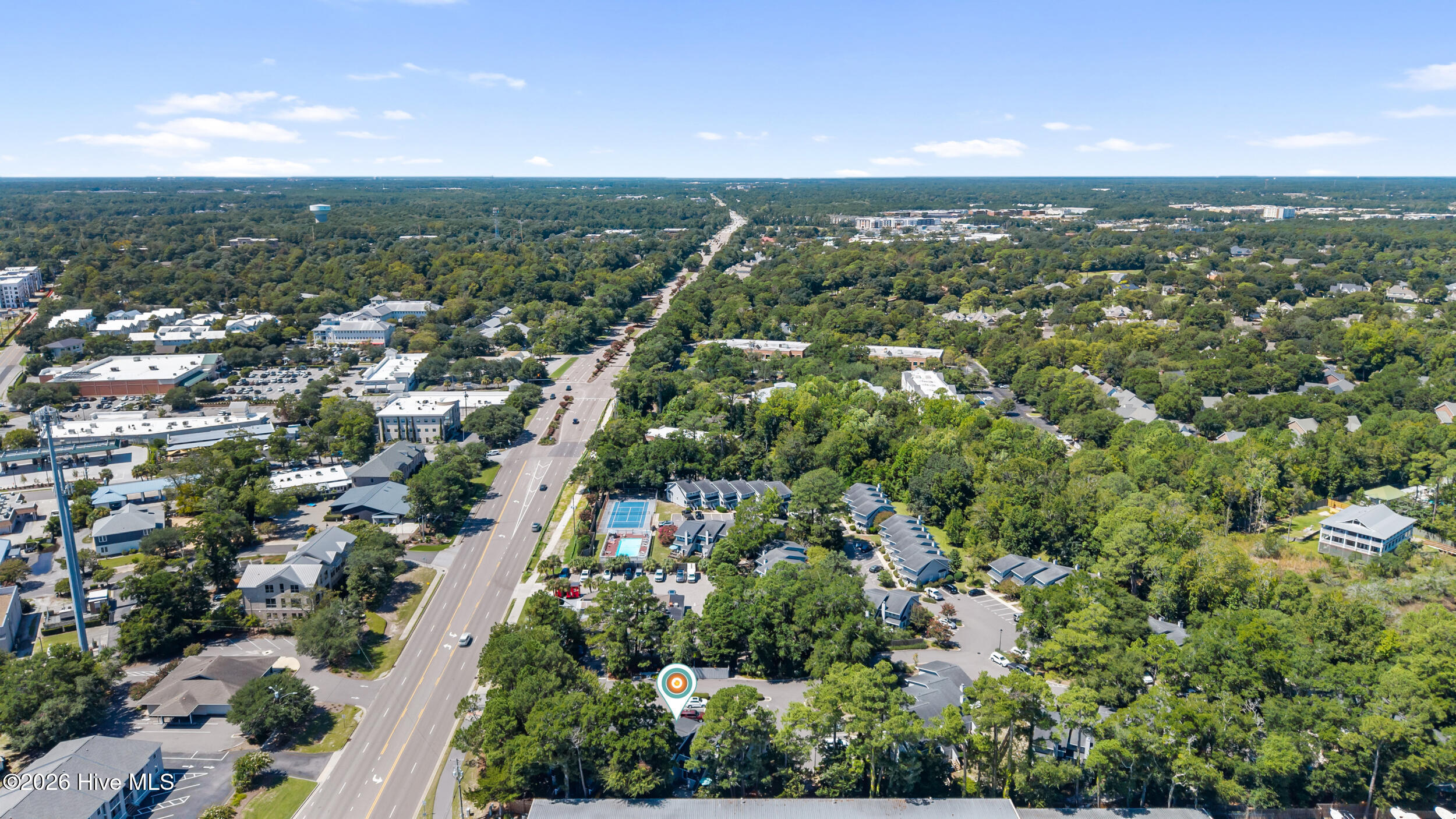 Aerial view of a wooded neighborhood and main road, with tennis courts and a pool near the marked property.
