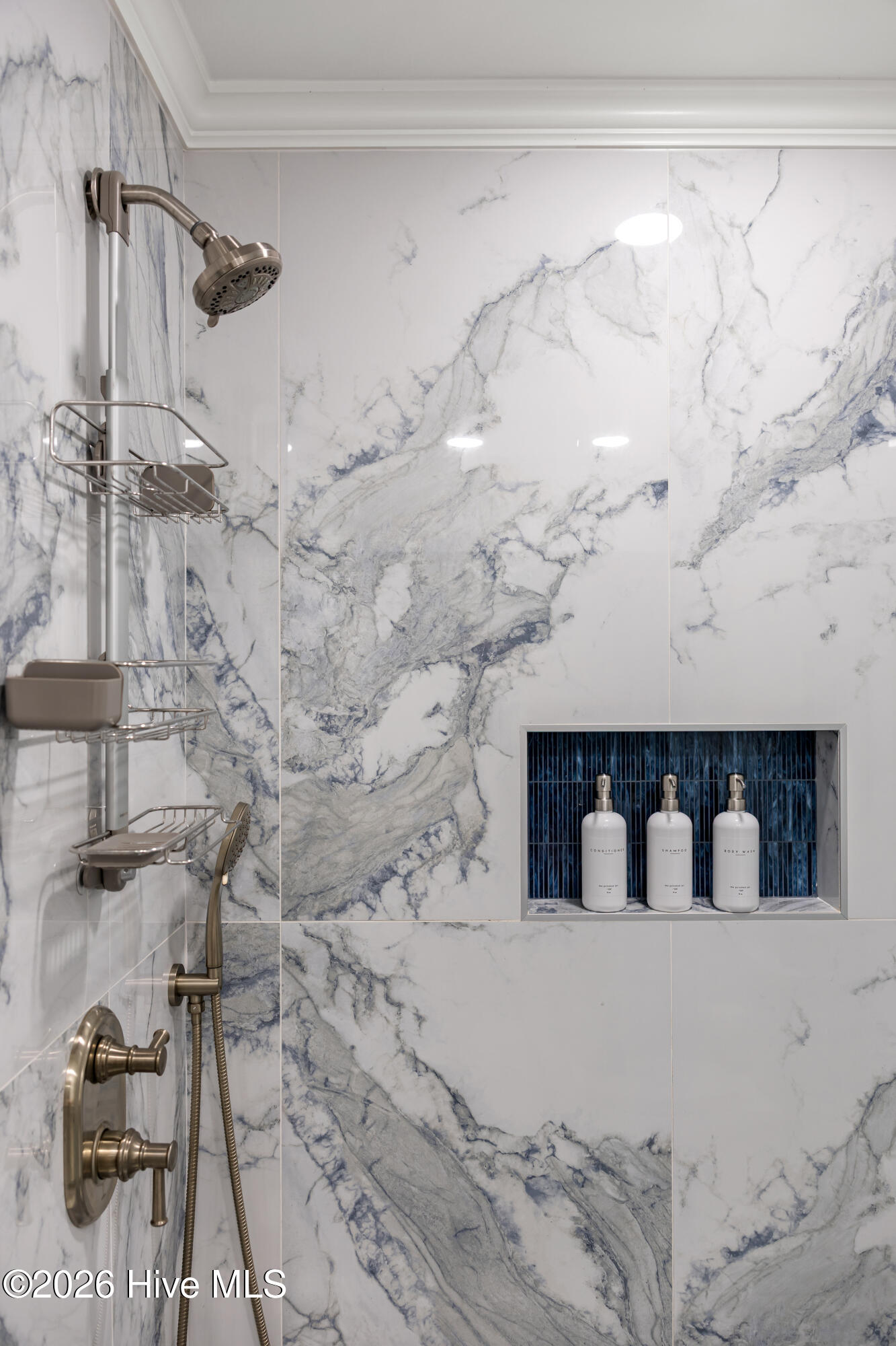 Marble-tiled shower with brushed metal fixtures and a blue-tiled niche holding three bottles.