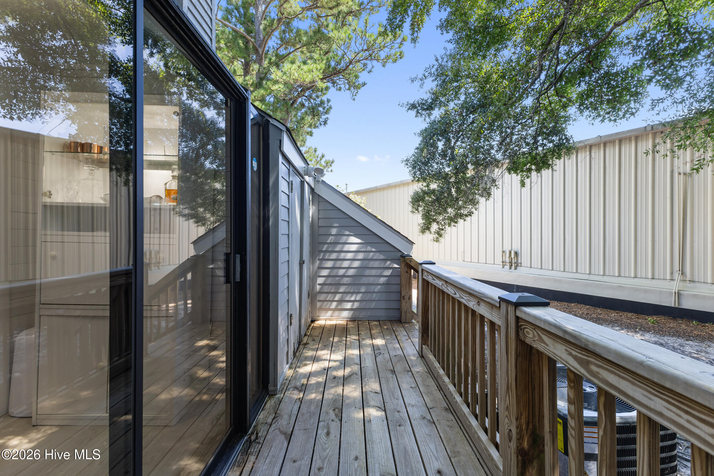 Raised wooden deck beside sliding glass doors, with a railing and leafy trees overhead.