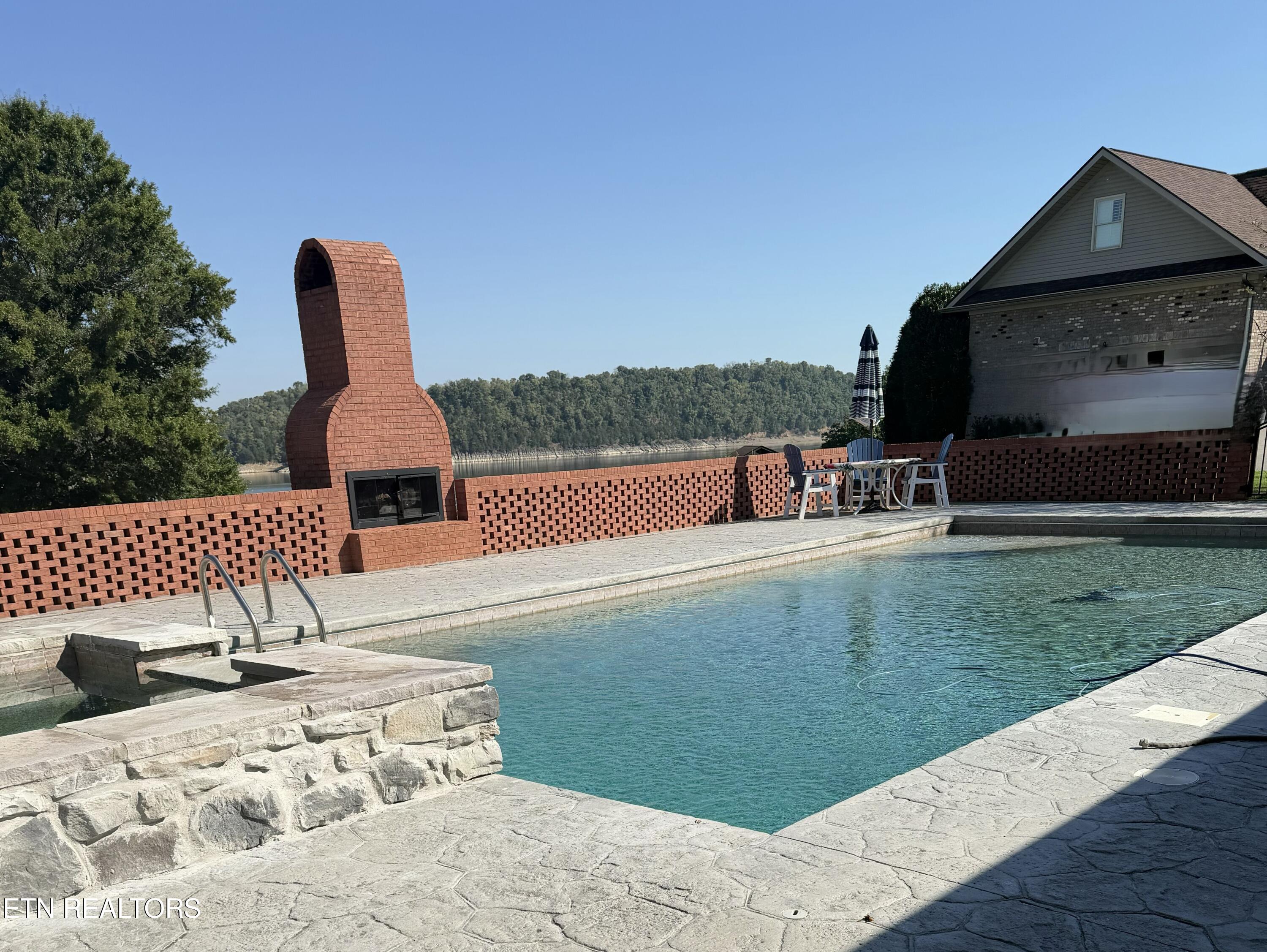 Swimming pool beside a stone patio, brick fireplace, and lake view.