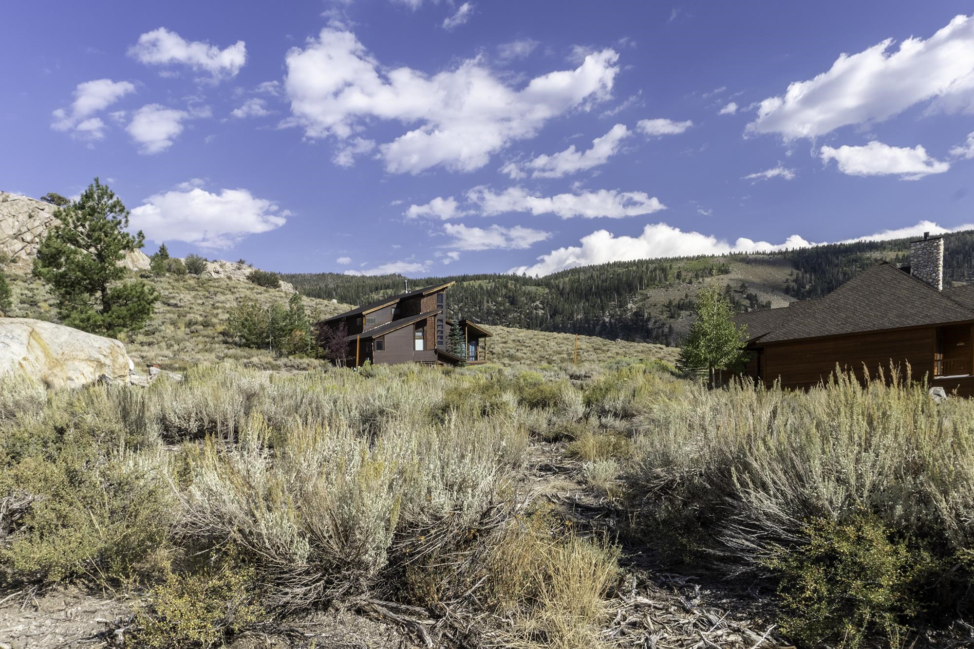 Wood mountain homes among sagebrush, with rocky outcrops and forested hills behind them.