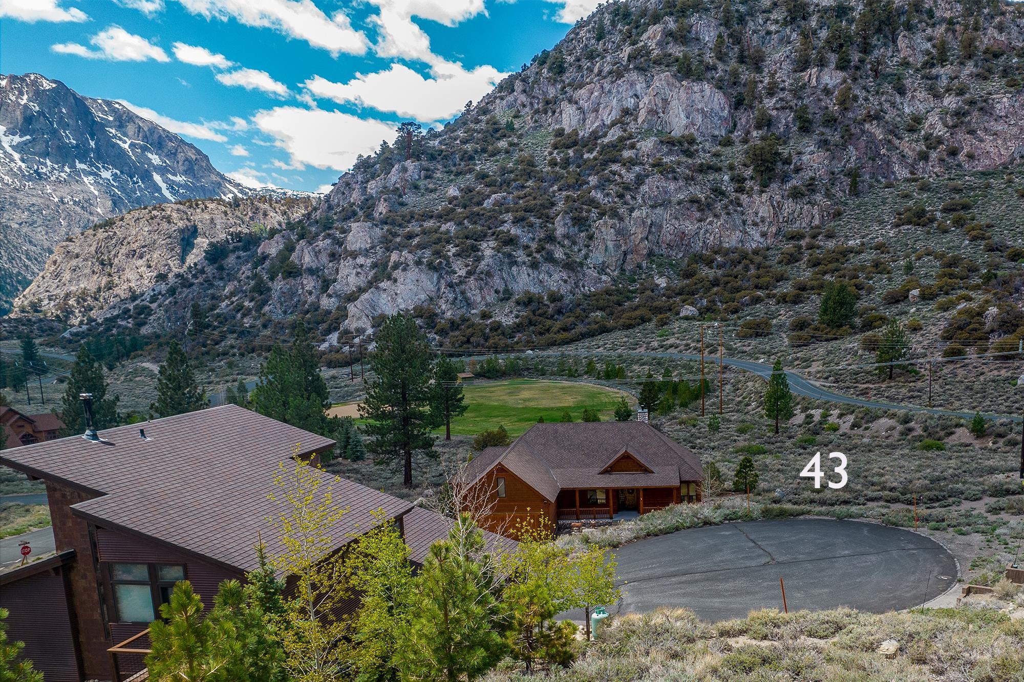 Brown cabin marked 43 below steep rocky mountains, with a paved driveway and neighboring homes.