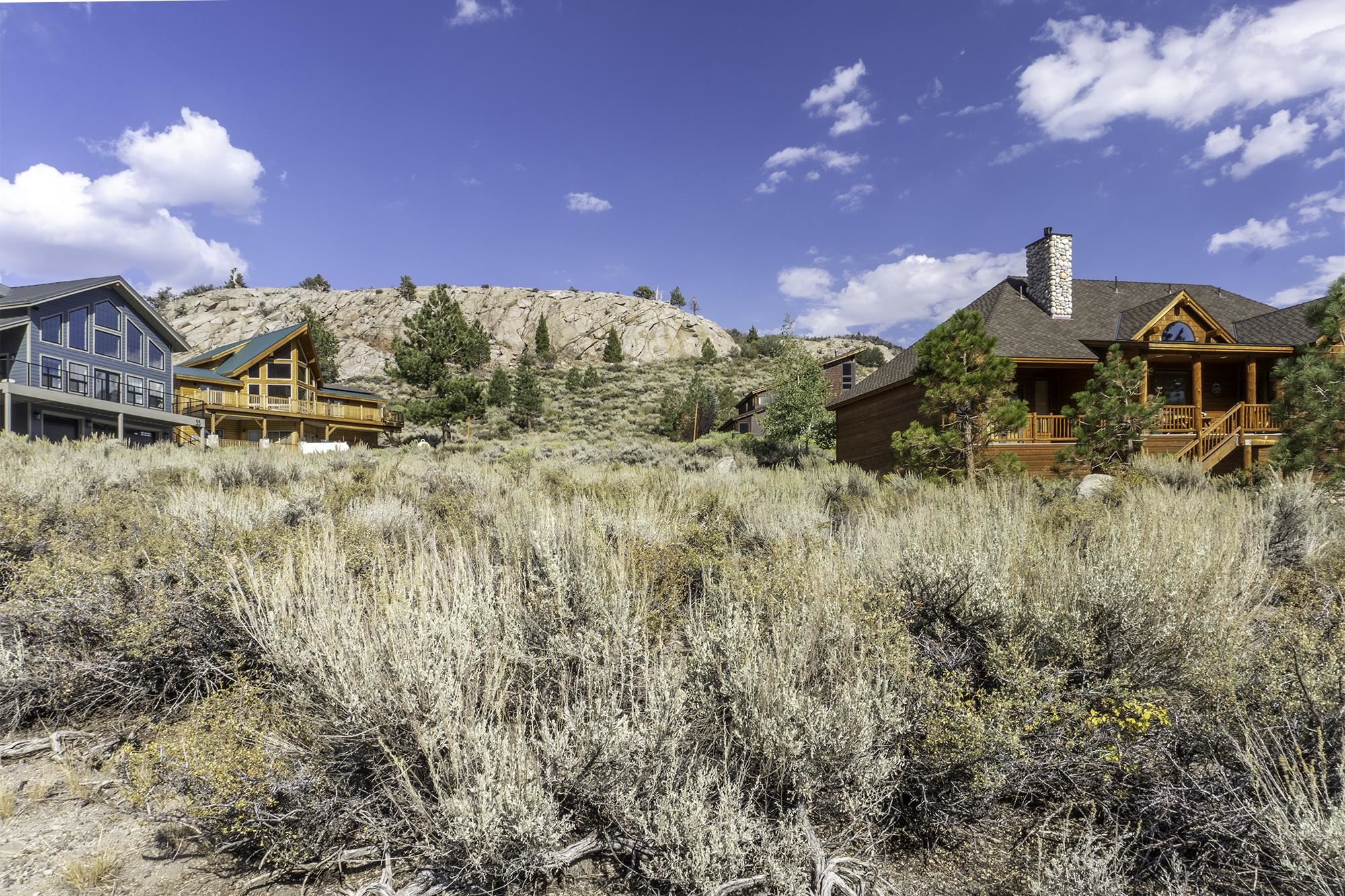 Wooden cabin with a broad porch and stone chimney among sagebrush below a rocky hillside.