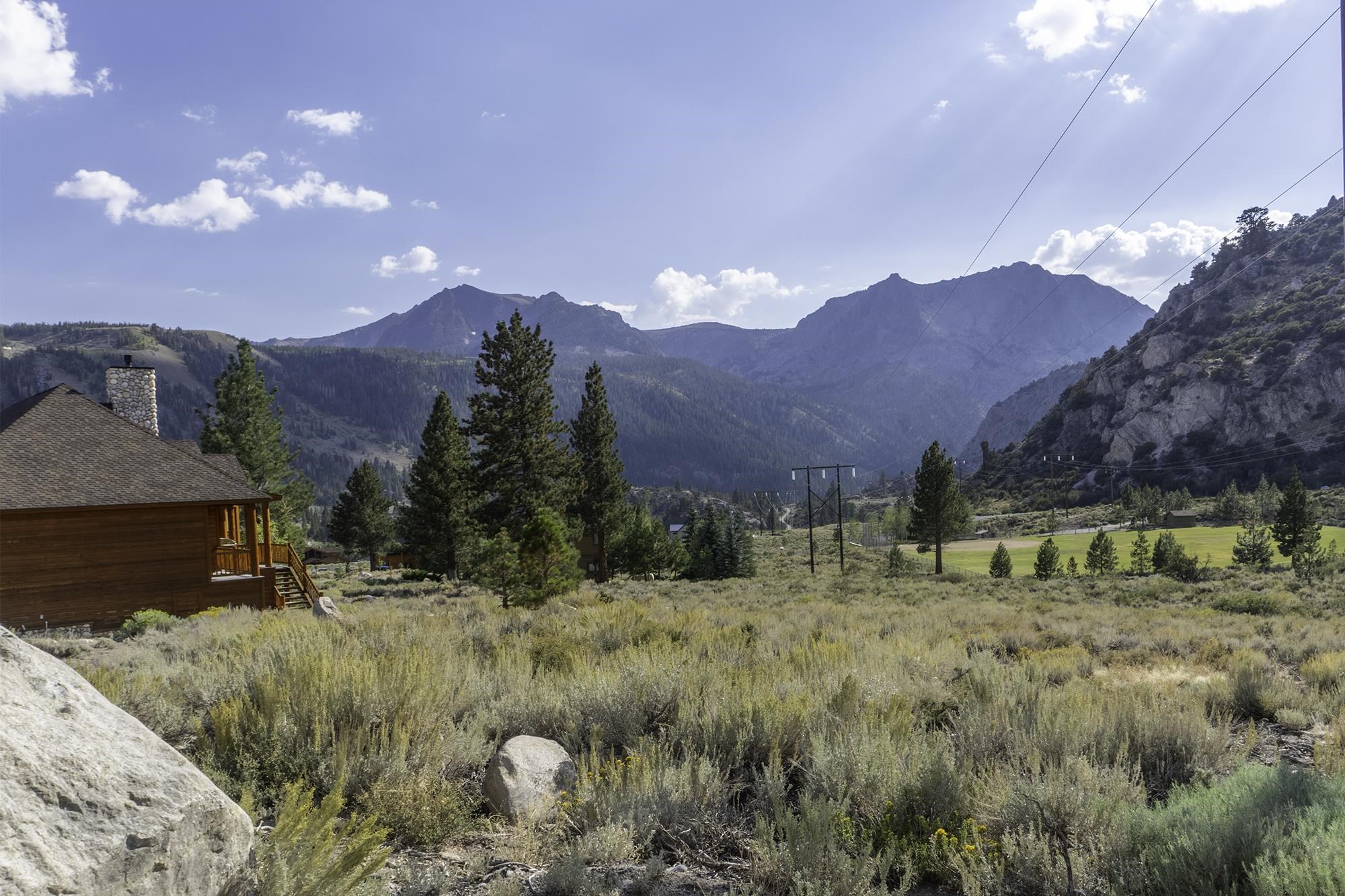 Mountain panorama beside a wood-sided cabin, with pine trees and a dry meadow in the foreground.