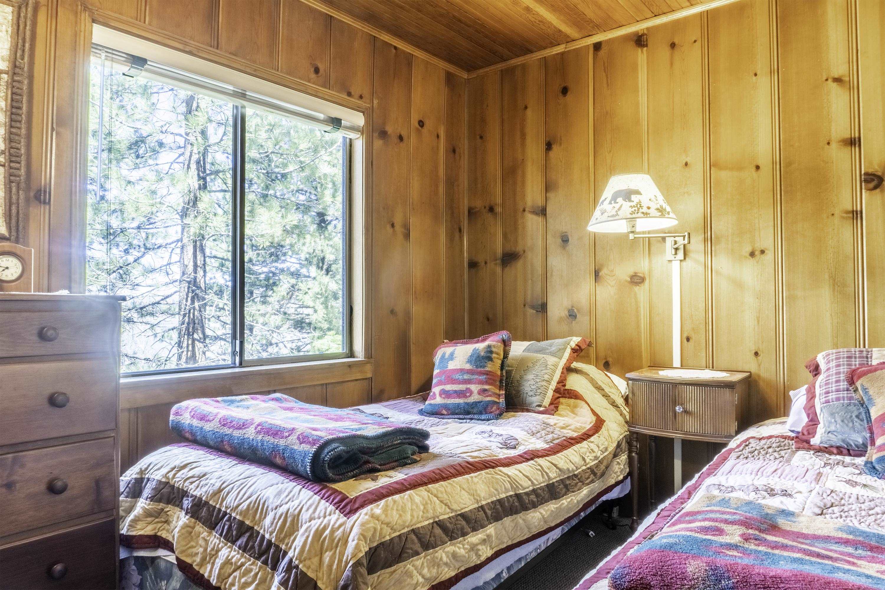 Bedroom with two quilt-covered beds, knotty pine walls, and a window overlooking pine trees.