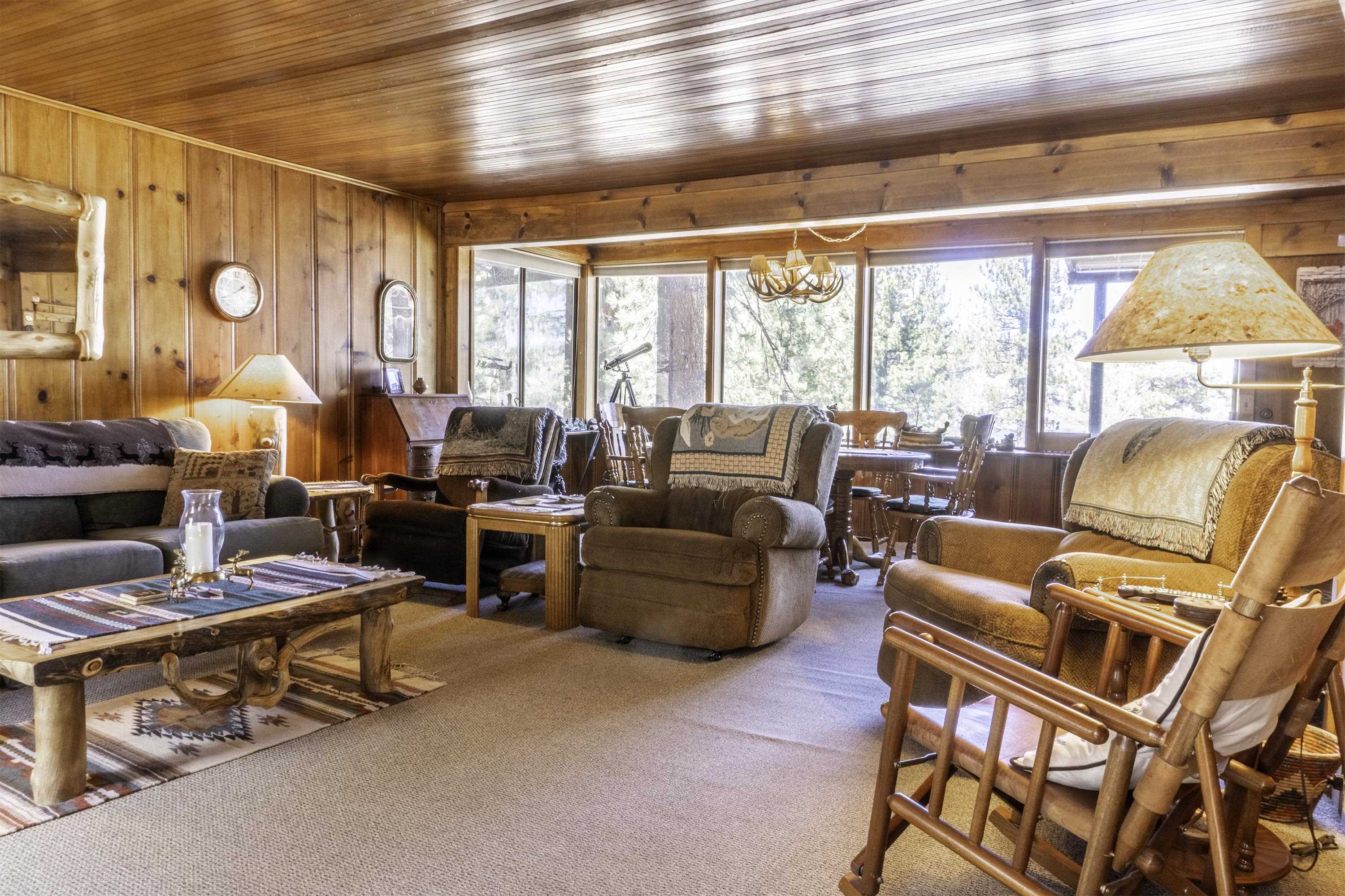 Living room with wood-paneled walls, cozy armchairs, and large windows overlooking trees.