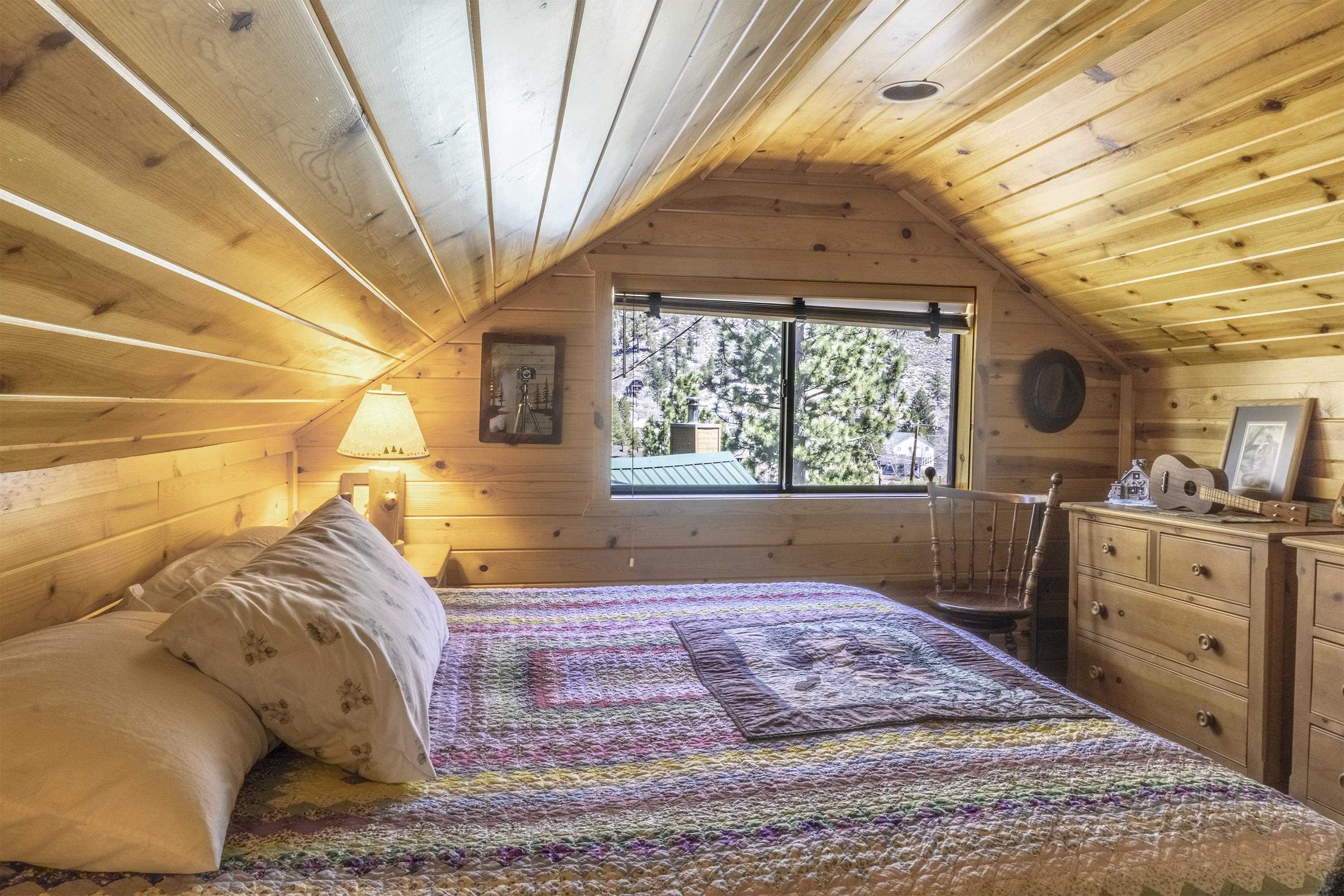 Attic bedroom with a colorful quilt beneath a sloped pine ceiling and a window overlooking trees.