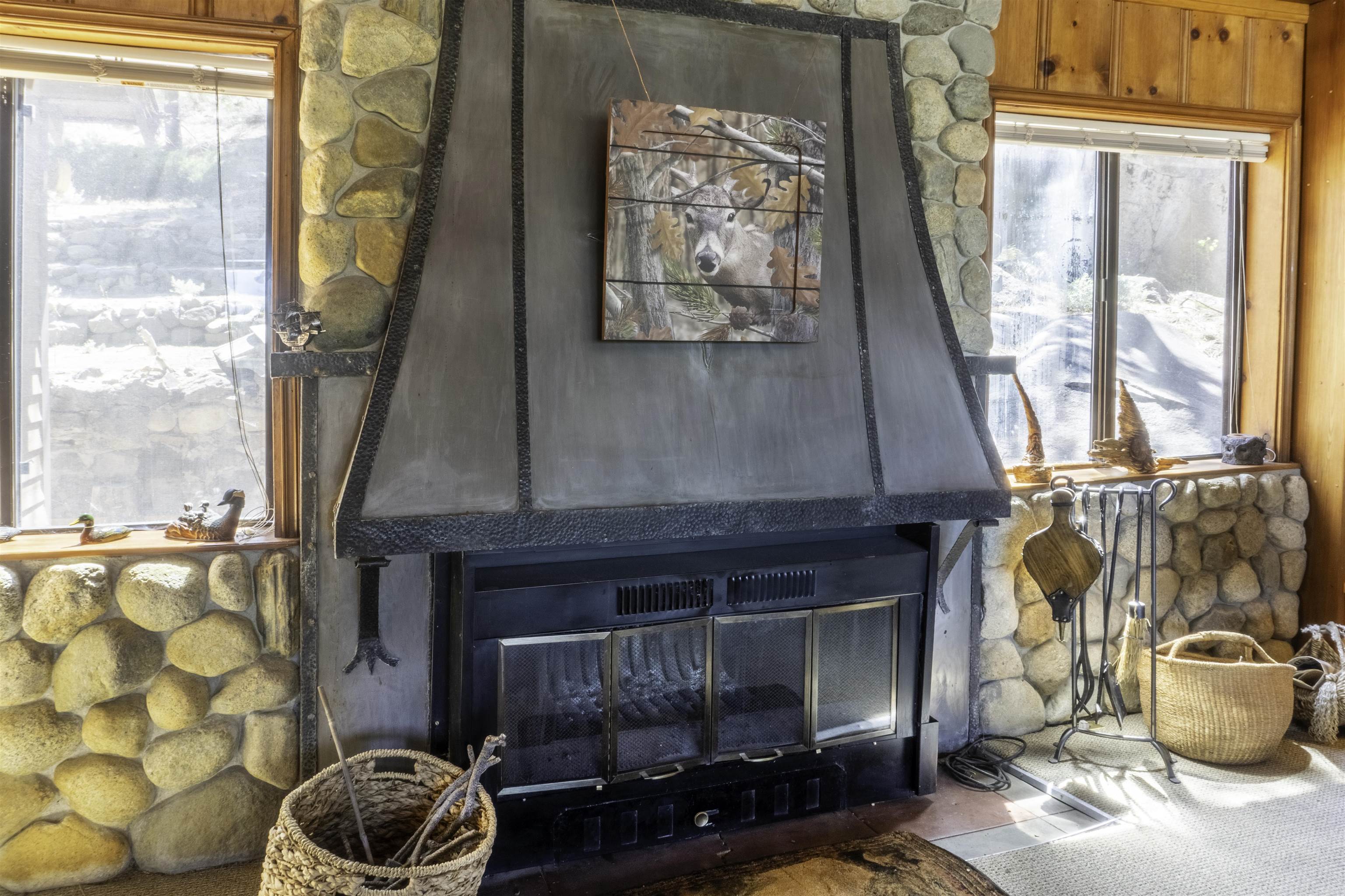 Stone fireplace with a black metal surround, deer artwork, and firewood baskets beside sunlit windows.