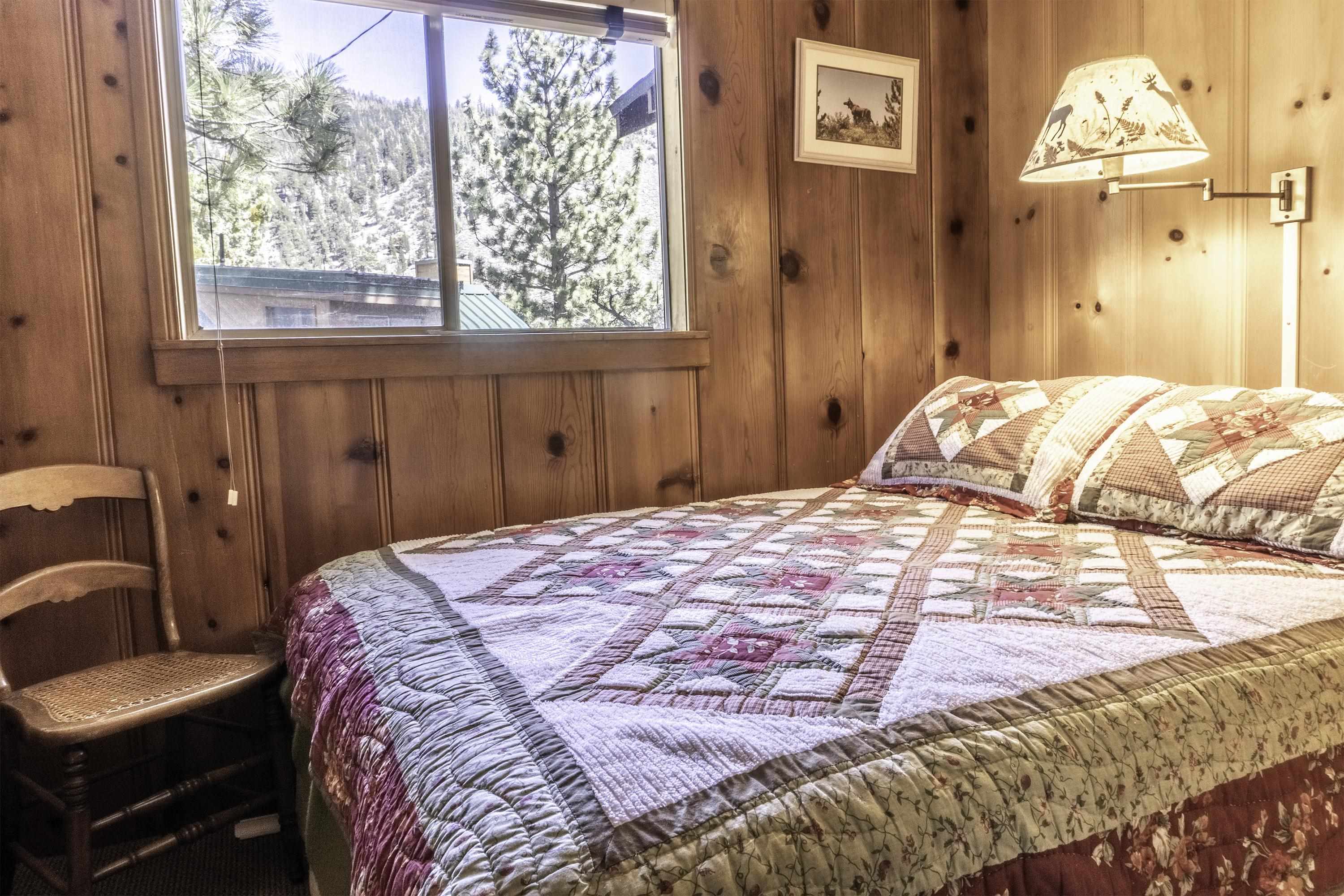 Bedroom with a patchwork quilt, knotty pine walls, and a window overlooking pine trees.