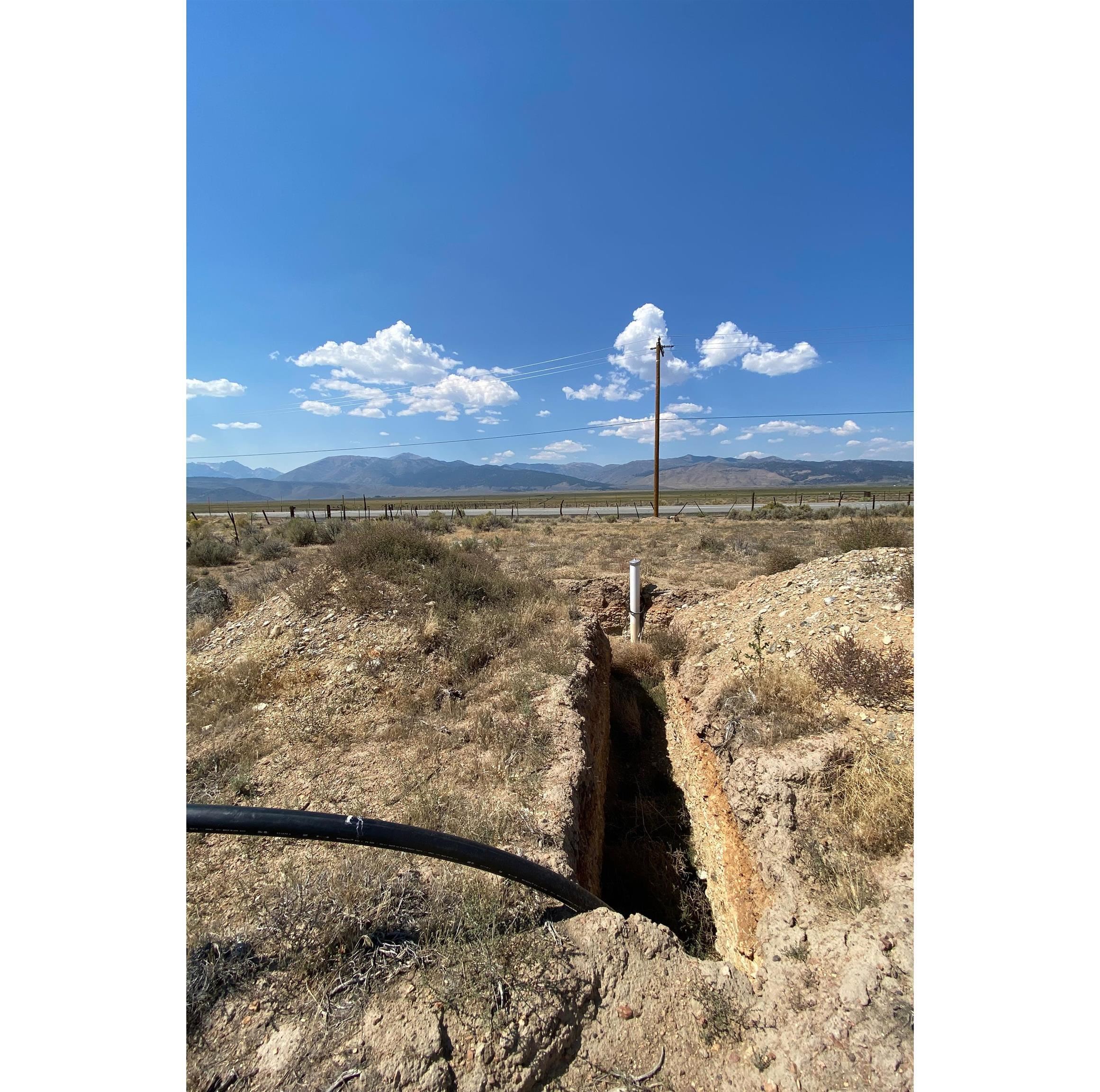 Dry vacant land with an open utility trench and pipe, overlooking a fenced valley and distant mountains.