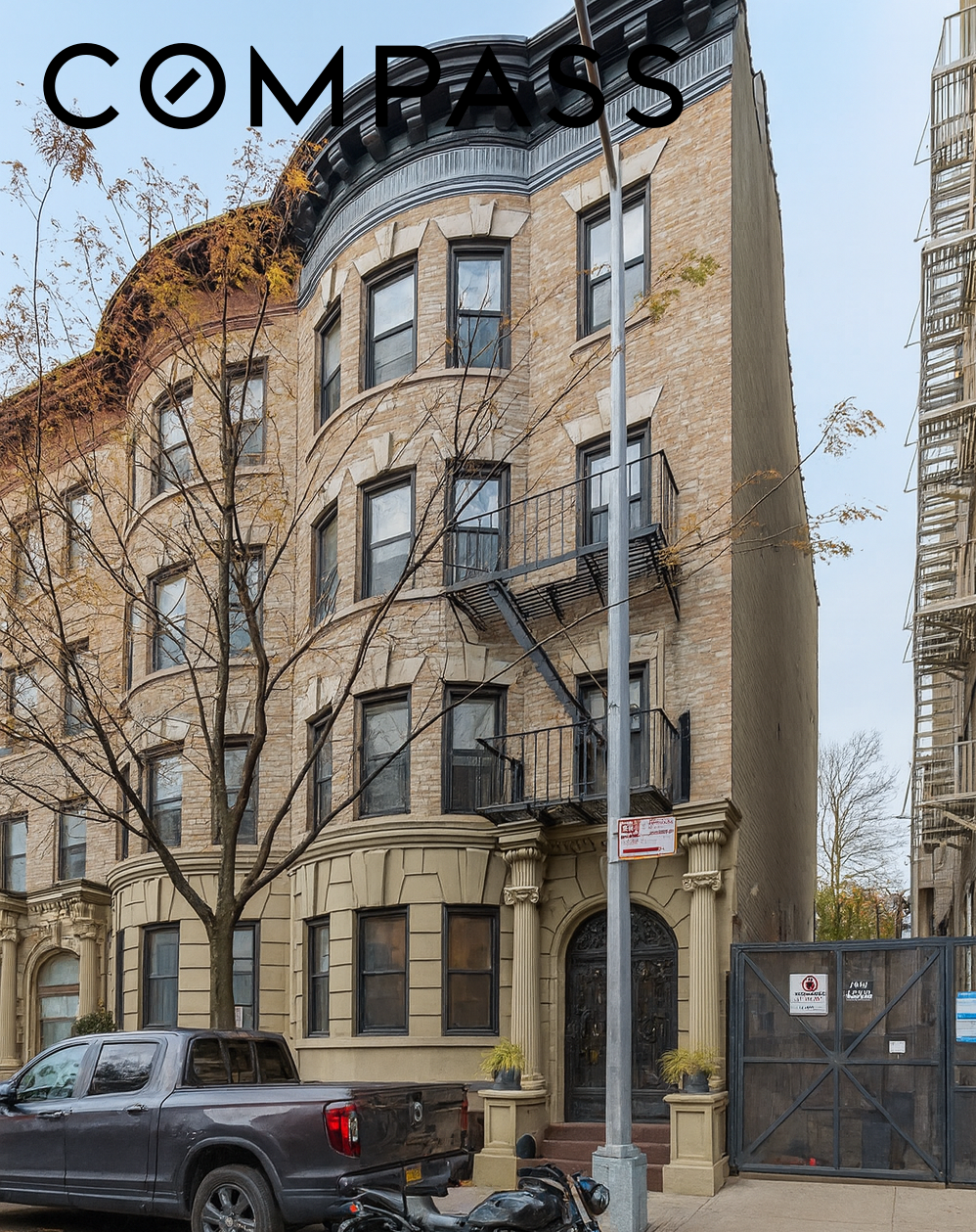 Tan brick residential building with rounded corner, balconies, and a leafless street tree.