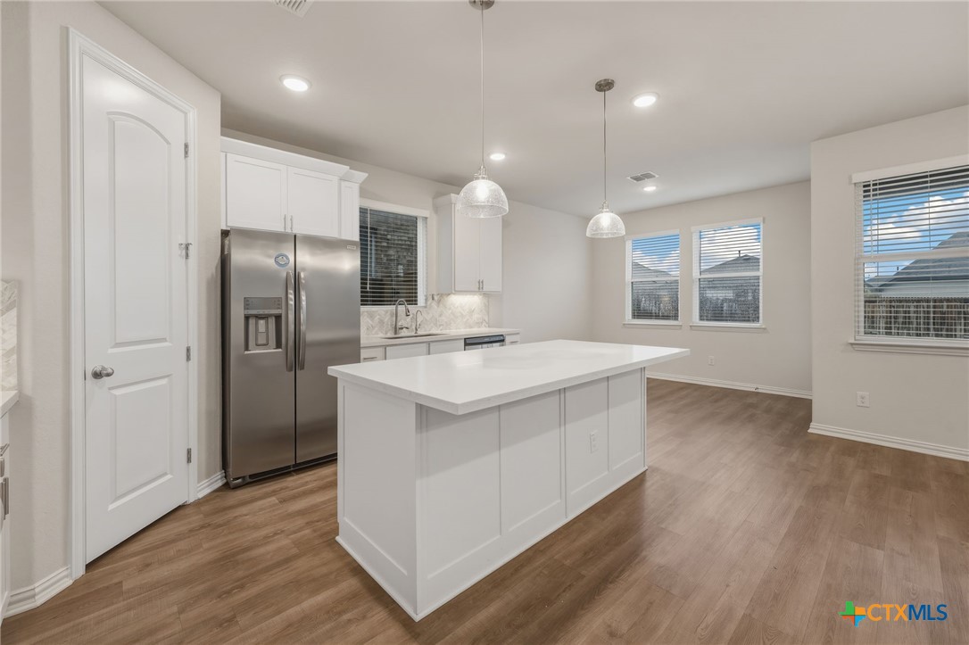 Kitchen with a large white island, stainless steel refrigerator, pendant lights, and wood-look flooring.