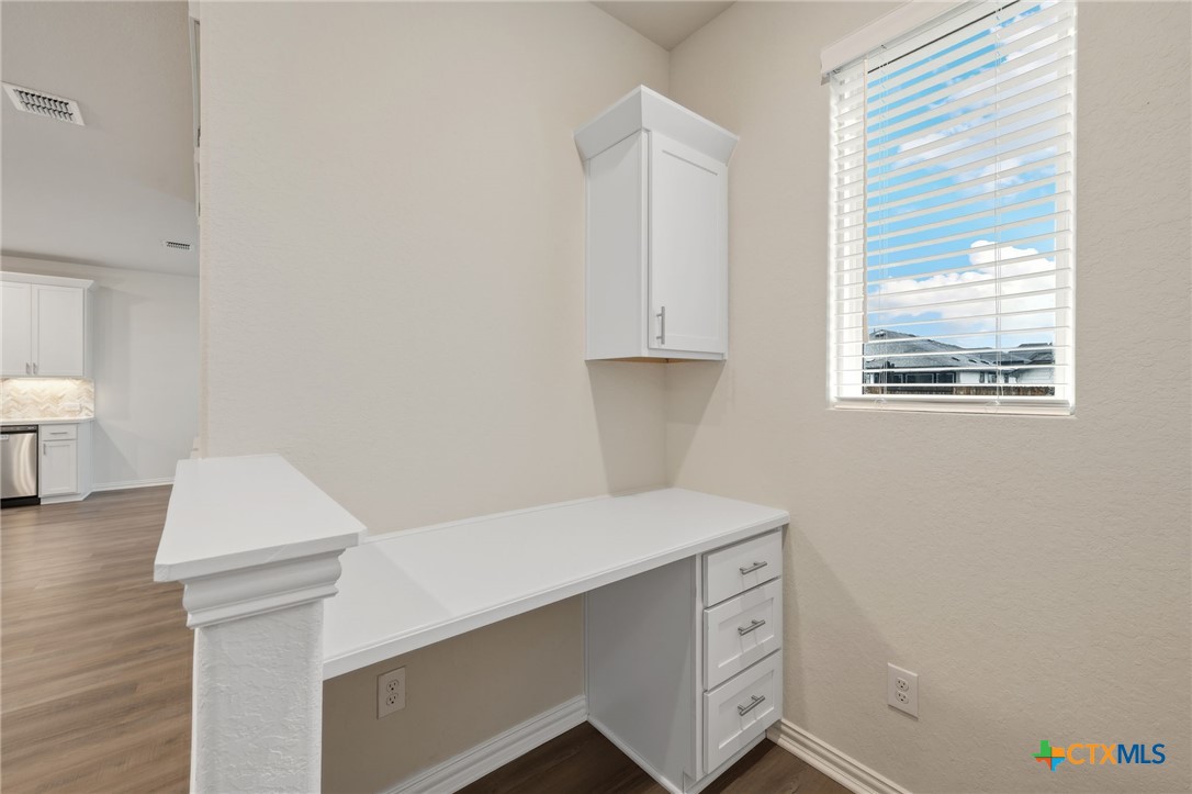 Built-in desk nook with white countertop, drawers, upper cabinet, and a window with blinds.