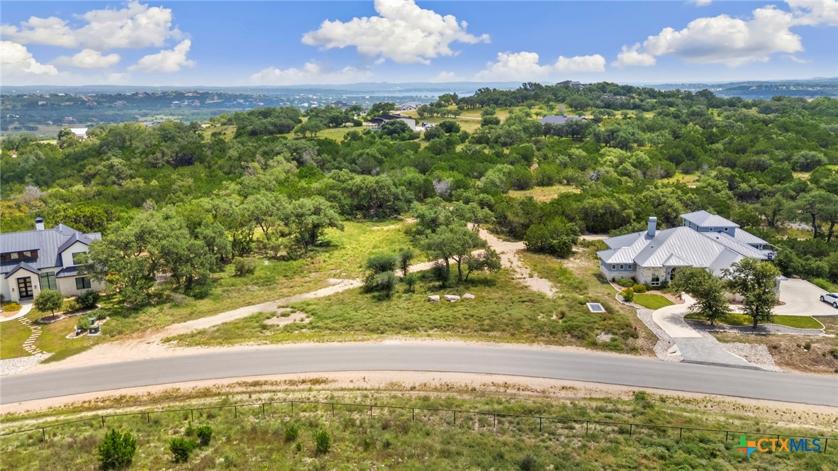 Vacant brush-covered parcel beside a curved road, between homes and rolling green hills.