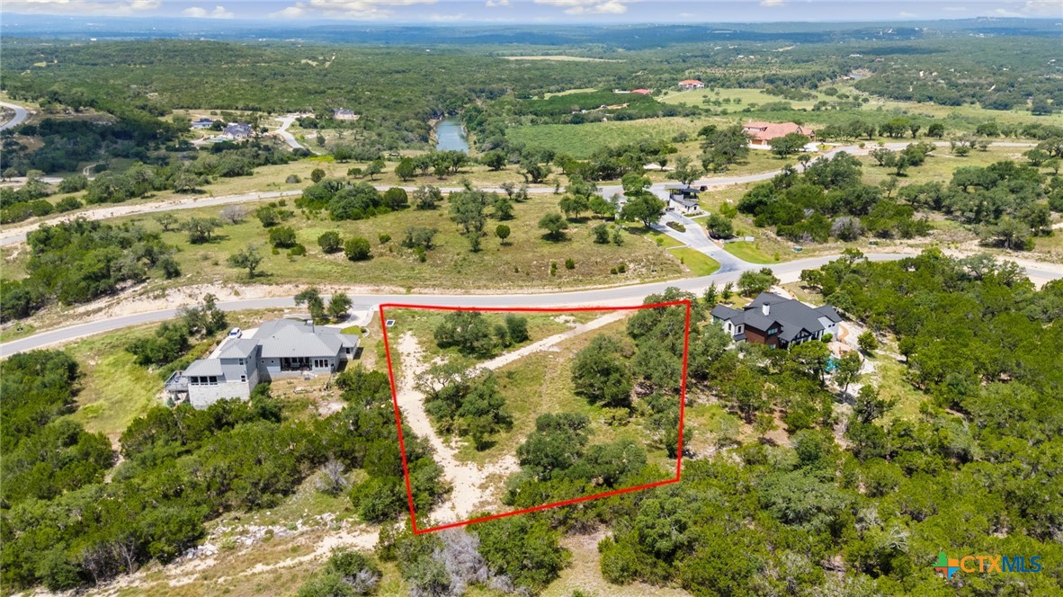 Aerial view of a brush-covered parcel outlined in red, with nearby homes and a small body of water in the distance.