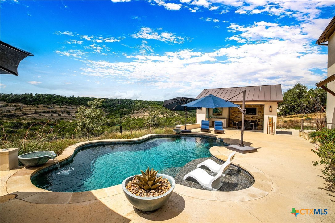 Curved swimming pool and sun shelf beside a covered outdoor kitchen, overlooking rolling hills.
