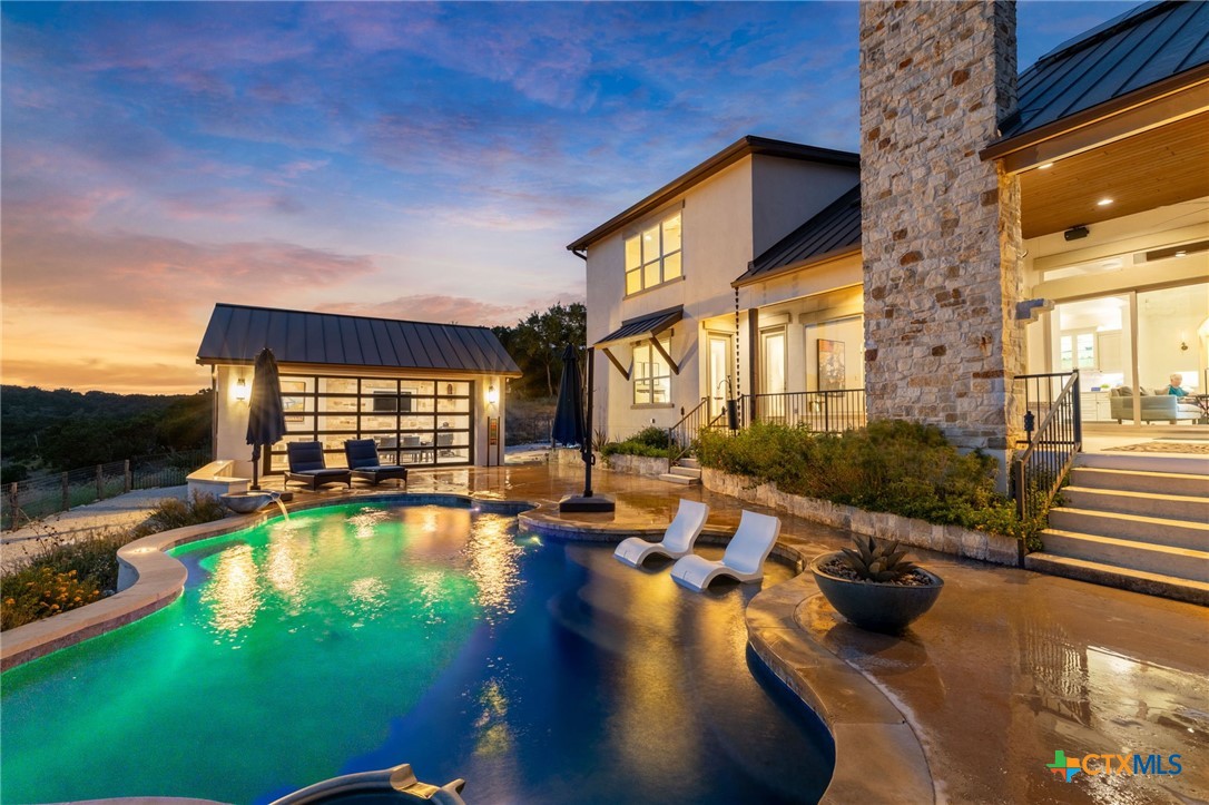 Illuminated swimming pool beside a modern home and detached pool house at dusk.