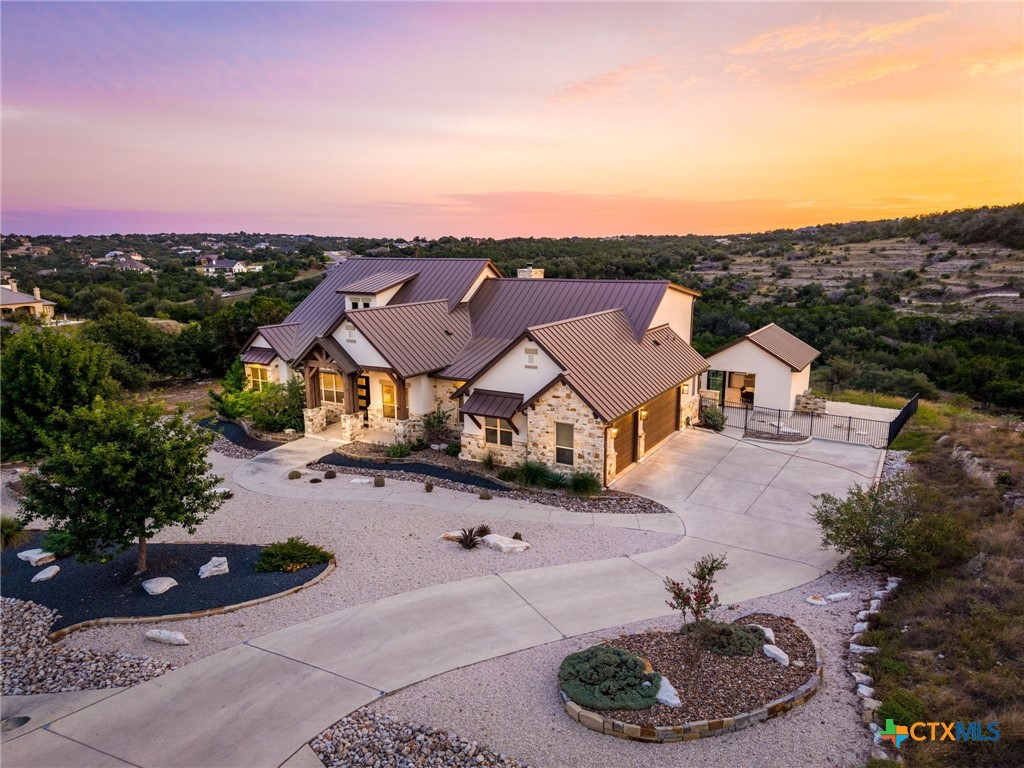 Aerial view of a stone-and-metal-roof home with a curved driveway at sunset.