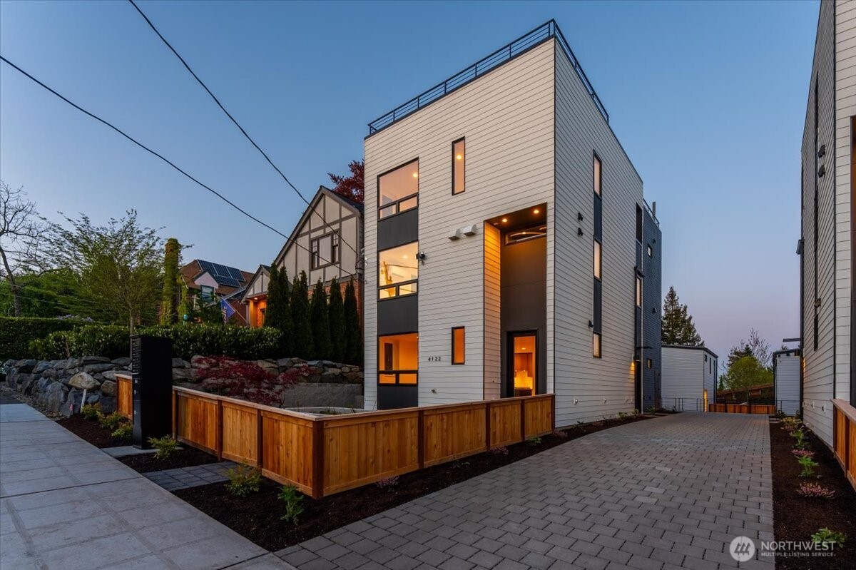 Modern multi-story Seattle home with light siding, a warmly lit entry, and a wooden front fence.