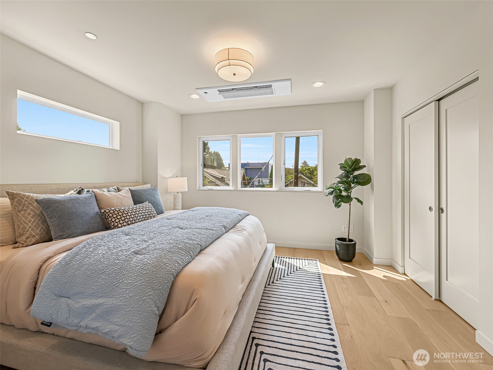 Staged bedroom with a large neutral bed, striped rug, and windows overlooking rooftops and trees.