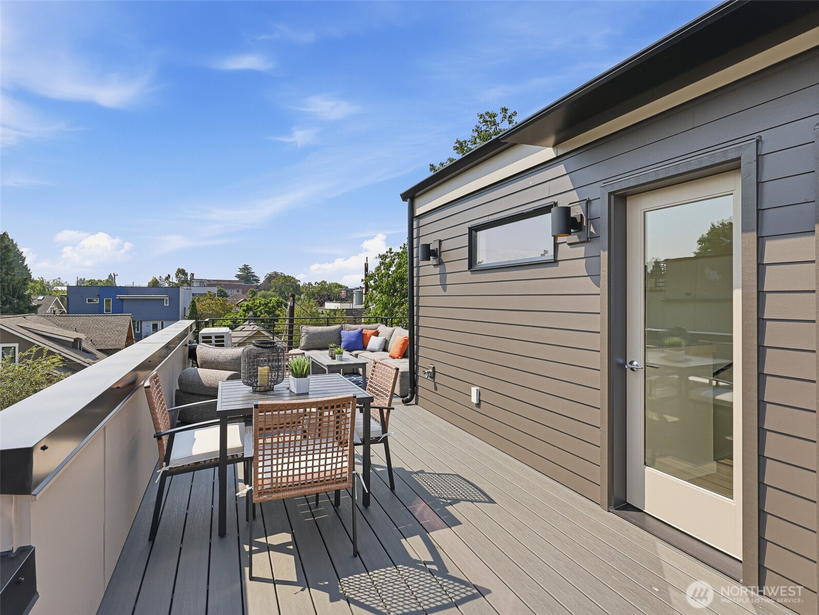Rooftop deck with outdoor dining set and cushioned seating overlooking nearby rooftops and trees.