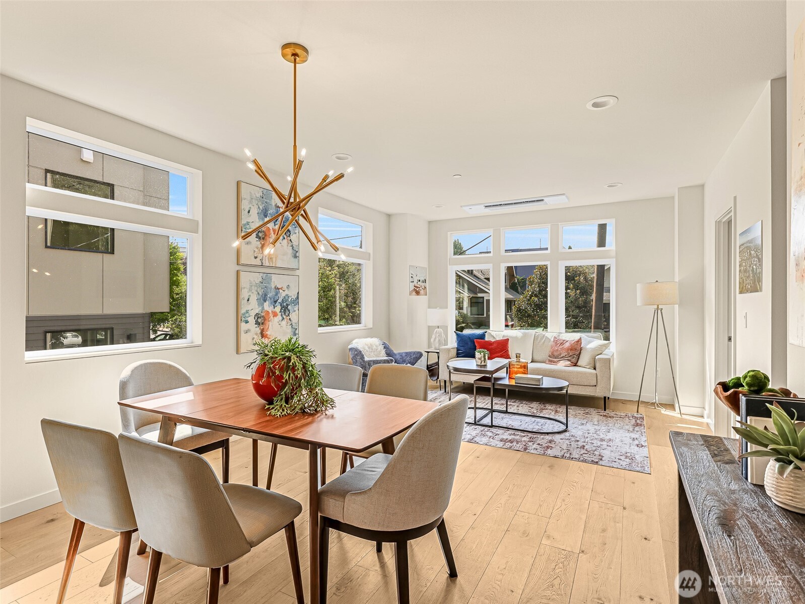 Open-plan living and dining room with light wood floors, large windows, and a brass chandelier.