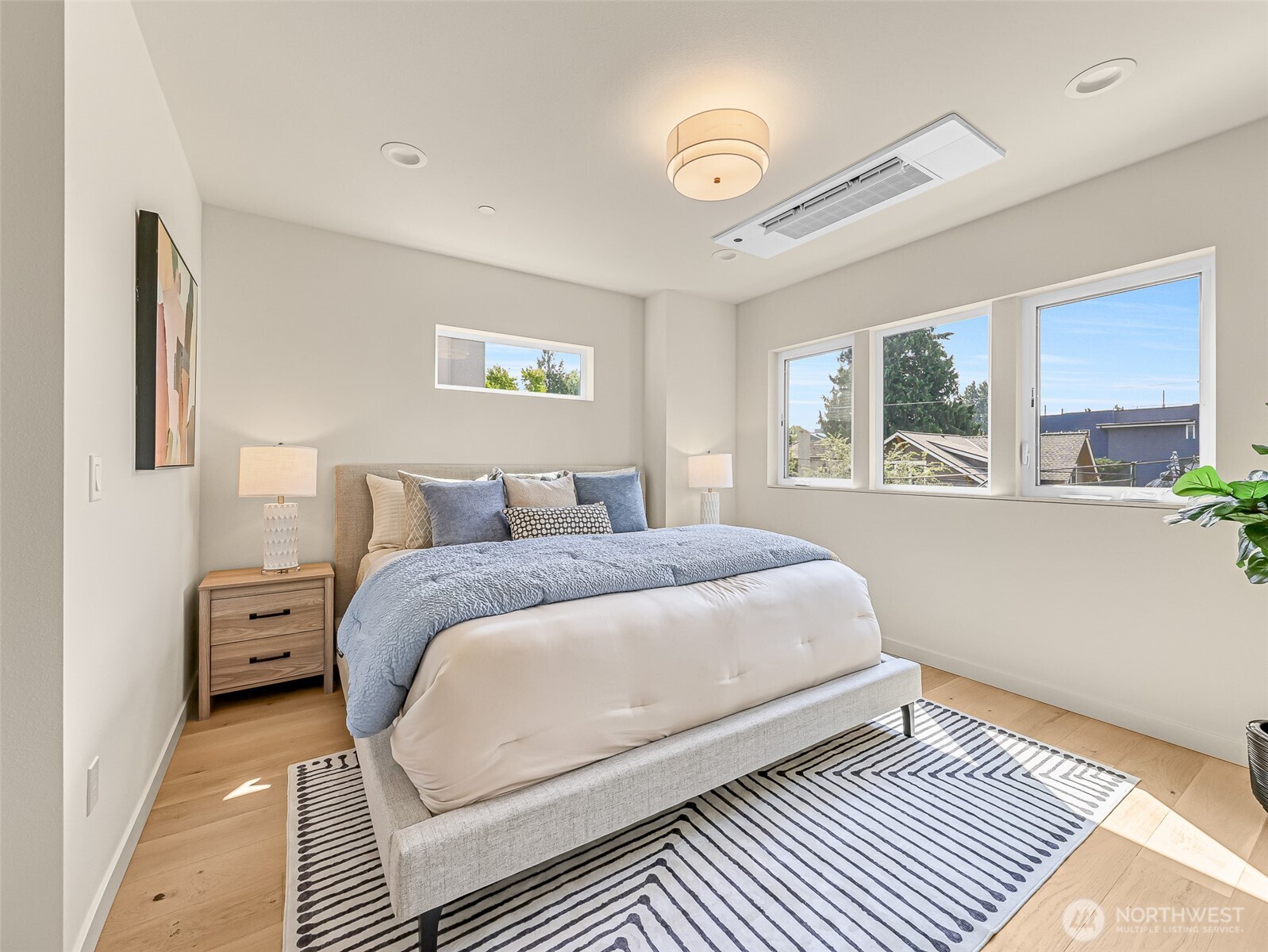 Furnished bedroom with a large bed, striped rug, and wide windows overlooking neighboring homes.