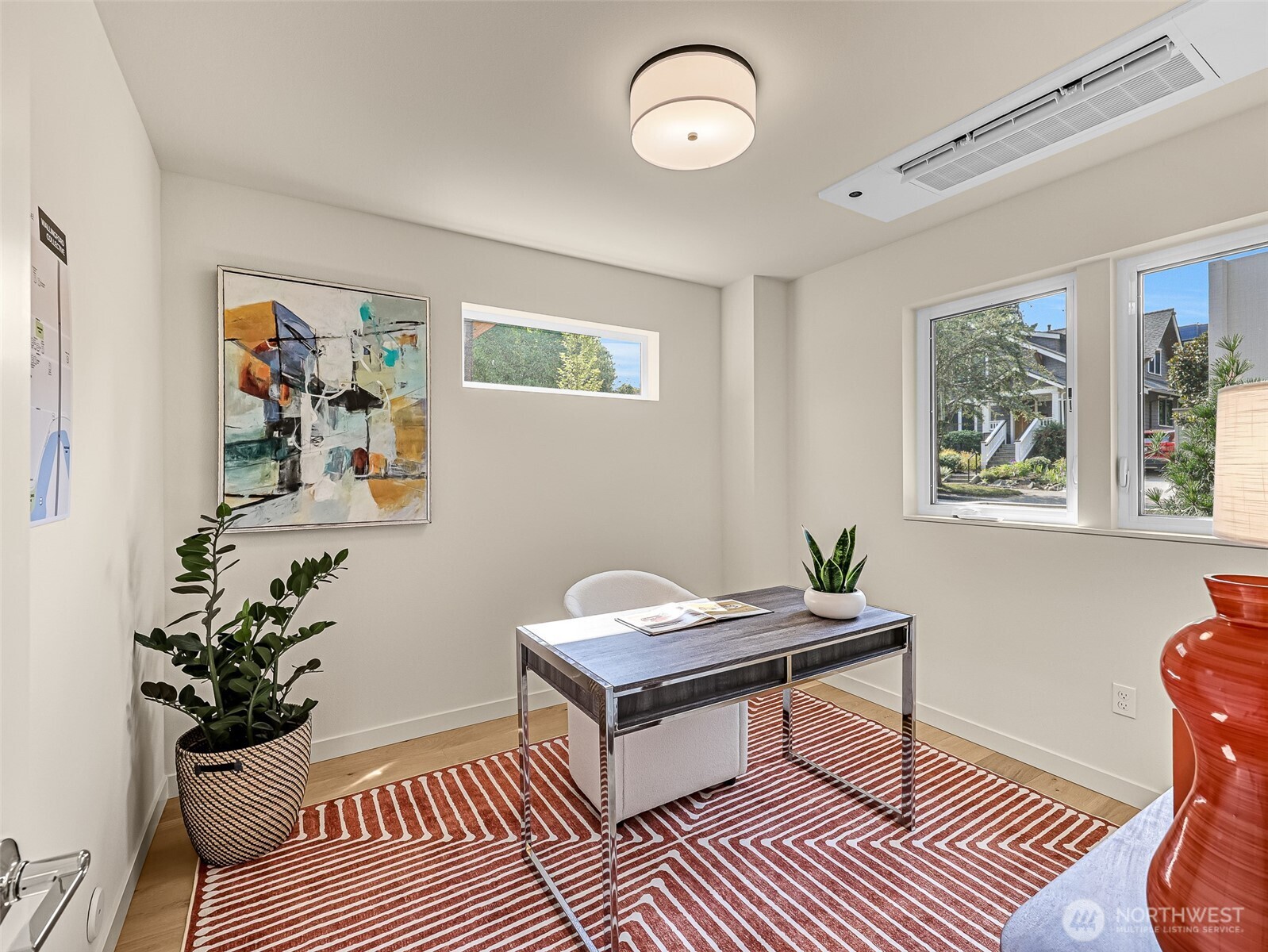 Furnished home office with a dark desk, abstract art, and windows overlooking a residential street.