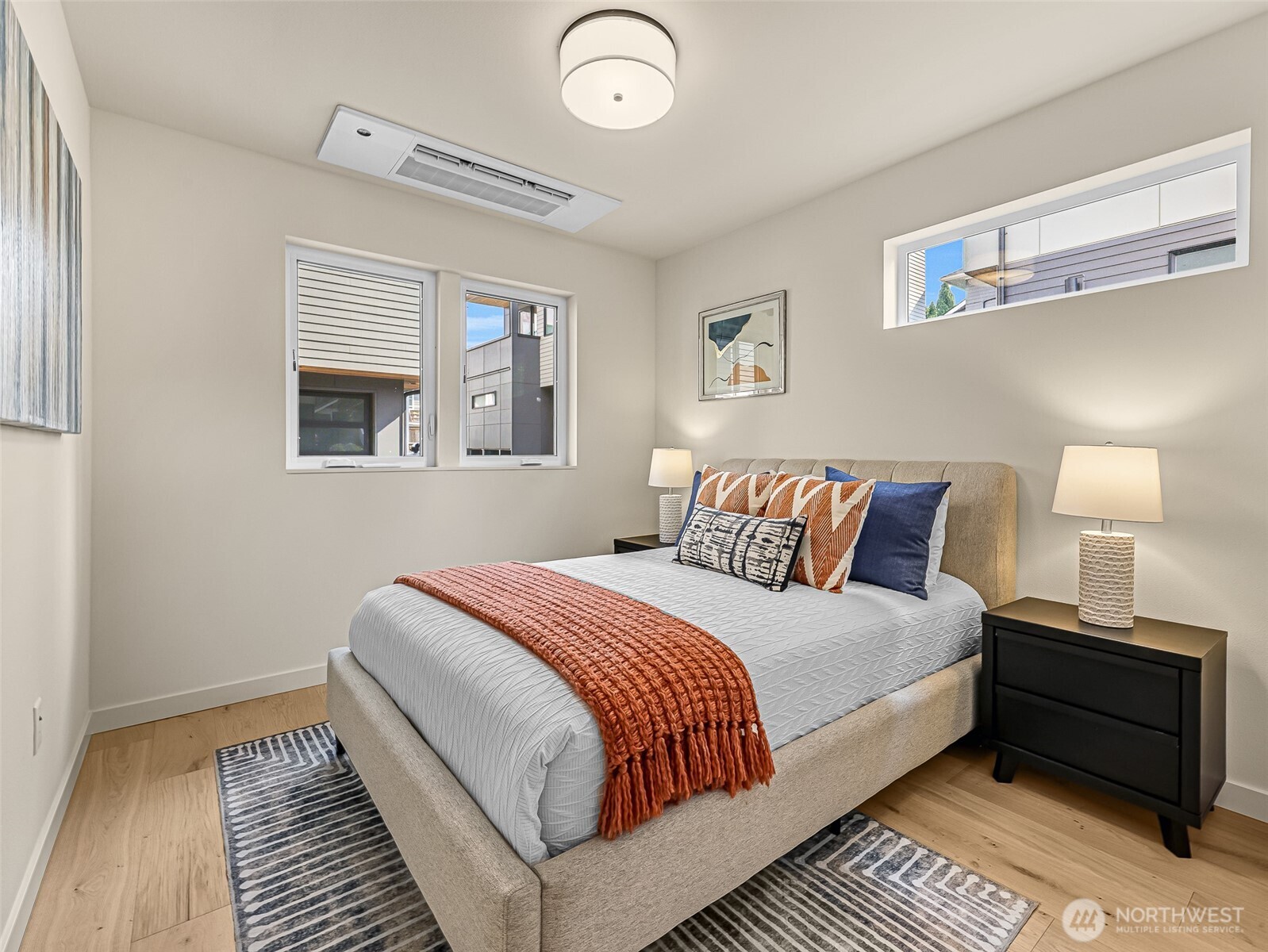 Staged bedroom with a queen-size bed, rust-colored throw, patterned rugs, and three windows.