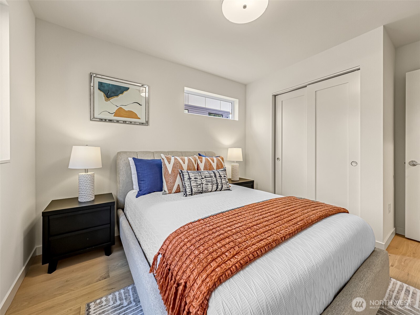 Staged bedroom with a gray bed, rust-colored throw, abstract art, and white sliding closet doors.
